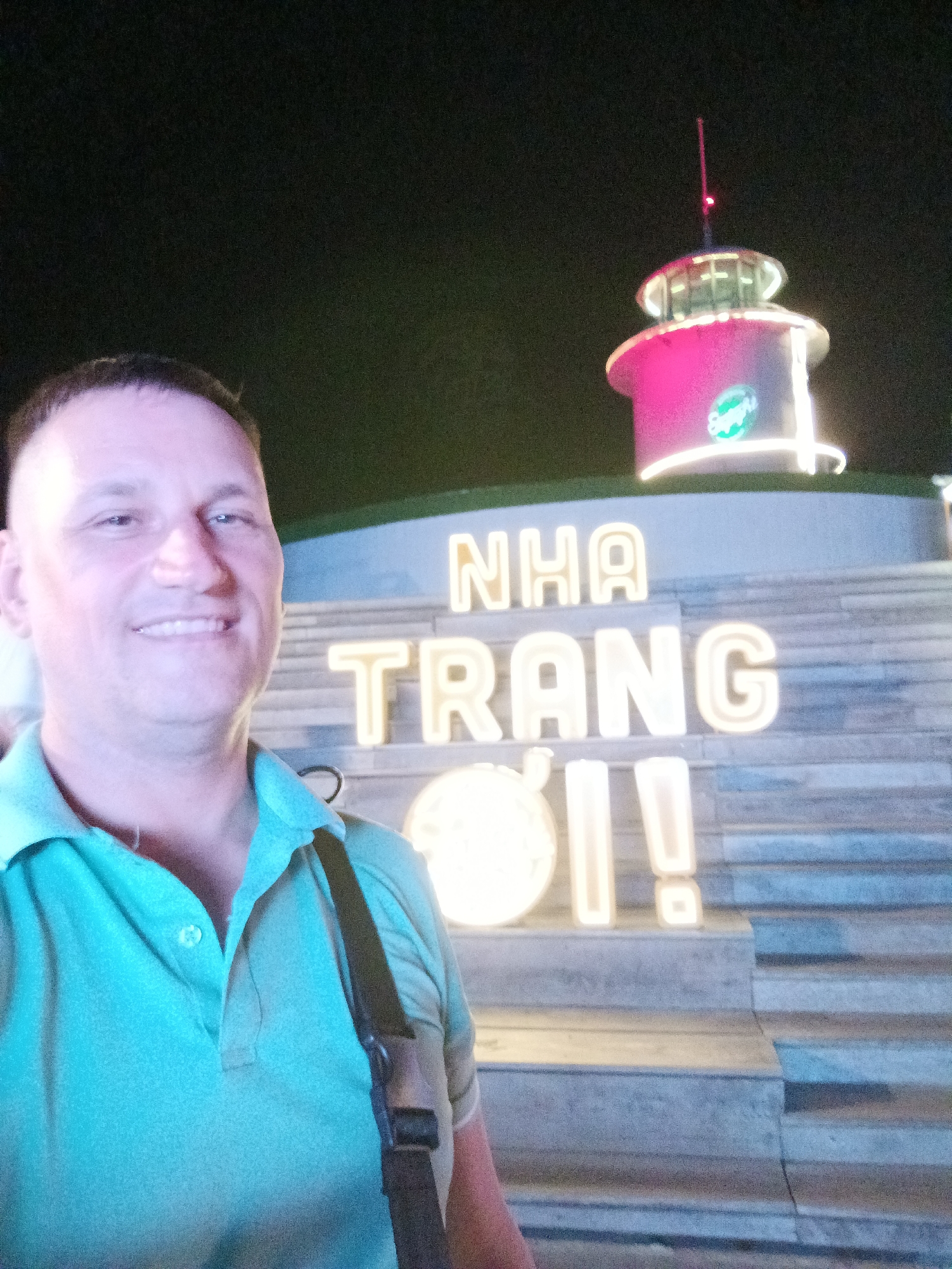 A medium shot depicts a man smiling at the camera, his face illuminated by ambient light. He is wearing a light turquoise polo shirt with a dark strap across his torso. Behind him, the illuminated words "NHA TRANG ƠI!" are displayed in large, capital letters on a tiered structure resembling a staircase. Above the lettering is a lit, circular building, possibly a small tower, with a red light on top. The background is dark, suggesting it is nighttime. The scene is in Nha Trang Ward, Vietnam.