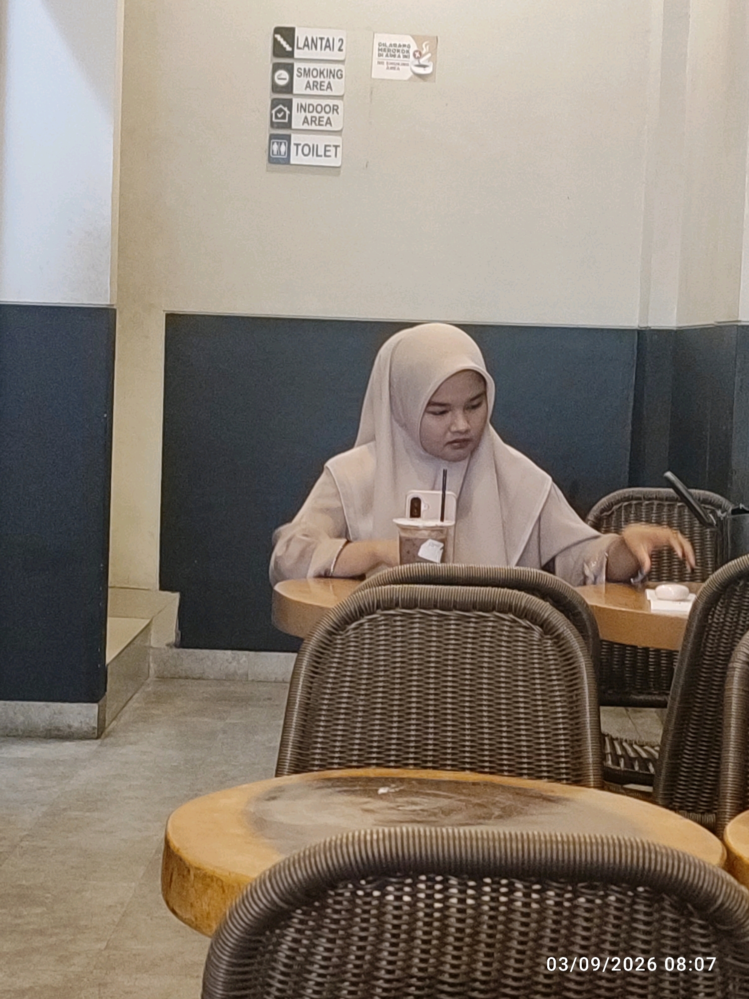 The image shows a woman wearing a light-colored hijab and an outfit of a similar shade, sitting at a table in what appears to be a cafe. She is looking down and to her right, with a cup on the table in front of her. The cafe has a neutral-colored wall with signage indicating "Lantai 2" (Floor 2), "Smoking Area," "Indoor Area," and "Toilet." Wicker chairs and wooden tables are visible. The timestamp "03/09/2026 08:07" is present in the lower right corner. This scene is set in a cafe in East Jakarta, Indonesia.