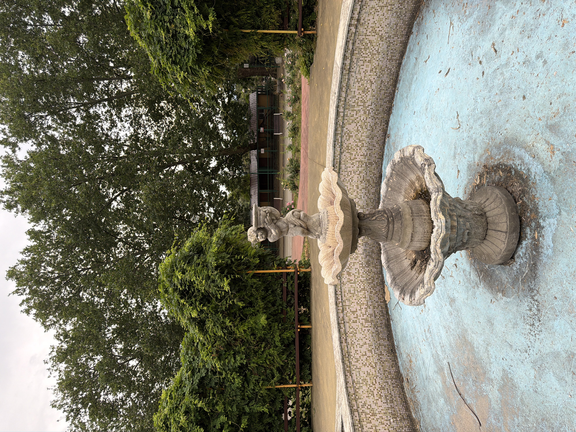 The image shows a dry, ornate stone fountain with a cherubic figure at the top, situated in a circular basin with a tiled rim and a light blue, weathered bottom. The fountain is surrounded by lush green foliage and tall trees, suggesting an outdoor park or garden setting. In the background, there are wooden benches and some buildings with pitched roofs, possibly part of a recreational area or zoo. The overall scene depicts a somewhat neglected, yet picturesque, outdoor public space. The context provided indicates this location is in Pazardzhik, Bulgaria.