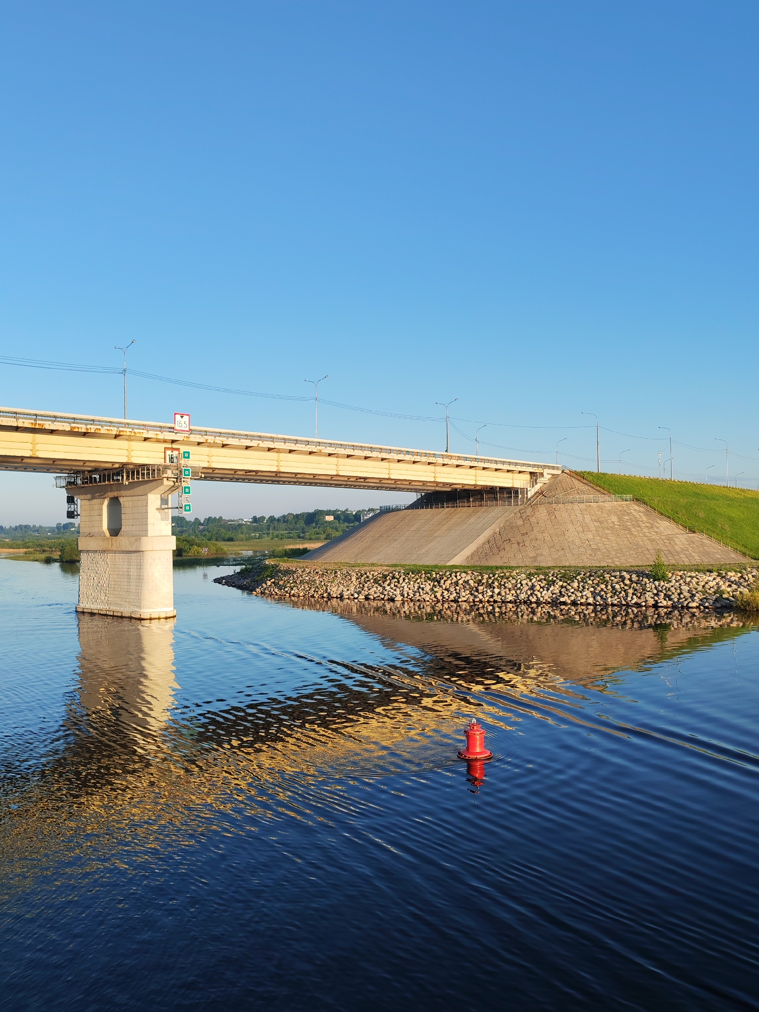 The image captures a bridge over water, likely a river or a wide canal. The bridge has a yellow-painted metal support structure and a concrete pier on one side. There are street lights along the bridge. A red buoy floats in the water, reflecting the bridge supports. The opposite bank features a large, grassy mound reinforced with rocks along the water's edge. The sky is clear and blue. The location is identified as Unknown, Unknown.