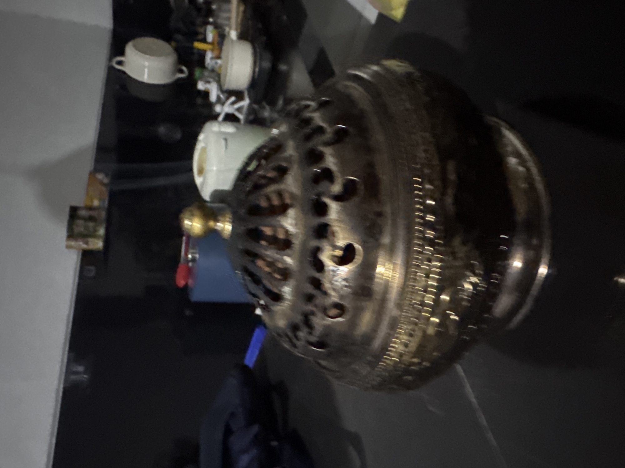 An ornate, ornate, ornate, brass, incense burner is displayed on a dark, reflective surface. The burner has a rounded, bulbous body with intricate carvings and a domed lid featuring an elaborate pattern of cutouts and a small, golden finial at the top. The background is blurred, but suggests a kitchen or countertop setting with shelves holding various items, including ceramic containers and bottles, hinting at a location in Sukawati, Indonesia.