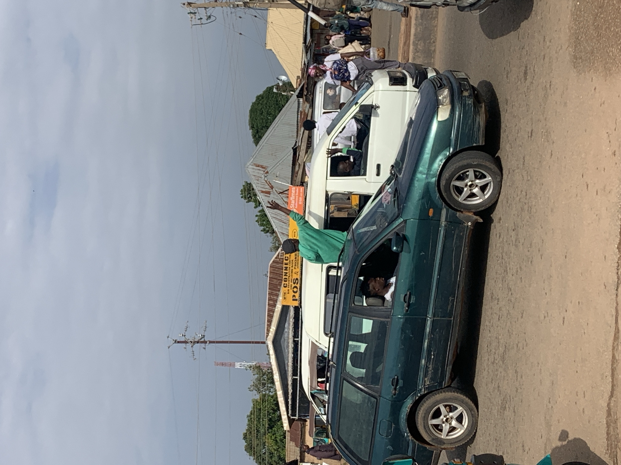 The image depicts a street scene in Gura Top, Nigeria, on a cloudy day. In the foreground, a dark green station wagon is parked. Inside the station wagon, a young Black man is visible, looking out the driver's side window. He is wearing a white tank top. A man in a bright green robe stands on top of a white van parked next to the station wagon, his right arm raised with his hand open. He appears to be speaking or gesturing. The white van has advertising signage on its side. Behind the white van, other vehicles and people are present, suggesting a busy urban environment. Buildings with corrugated metal roofs line the street, and utility poles with wires are visible in the background. The overall impression is of a lively, possibly commercial area in Gura Top.
