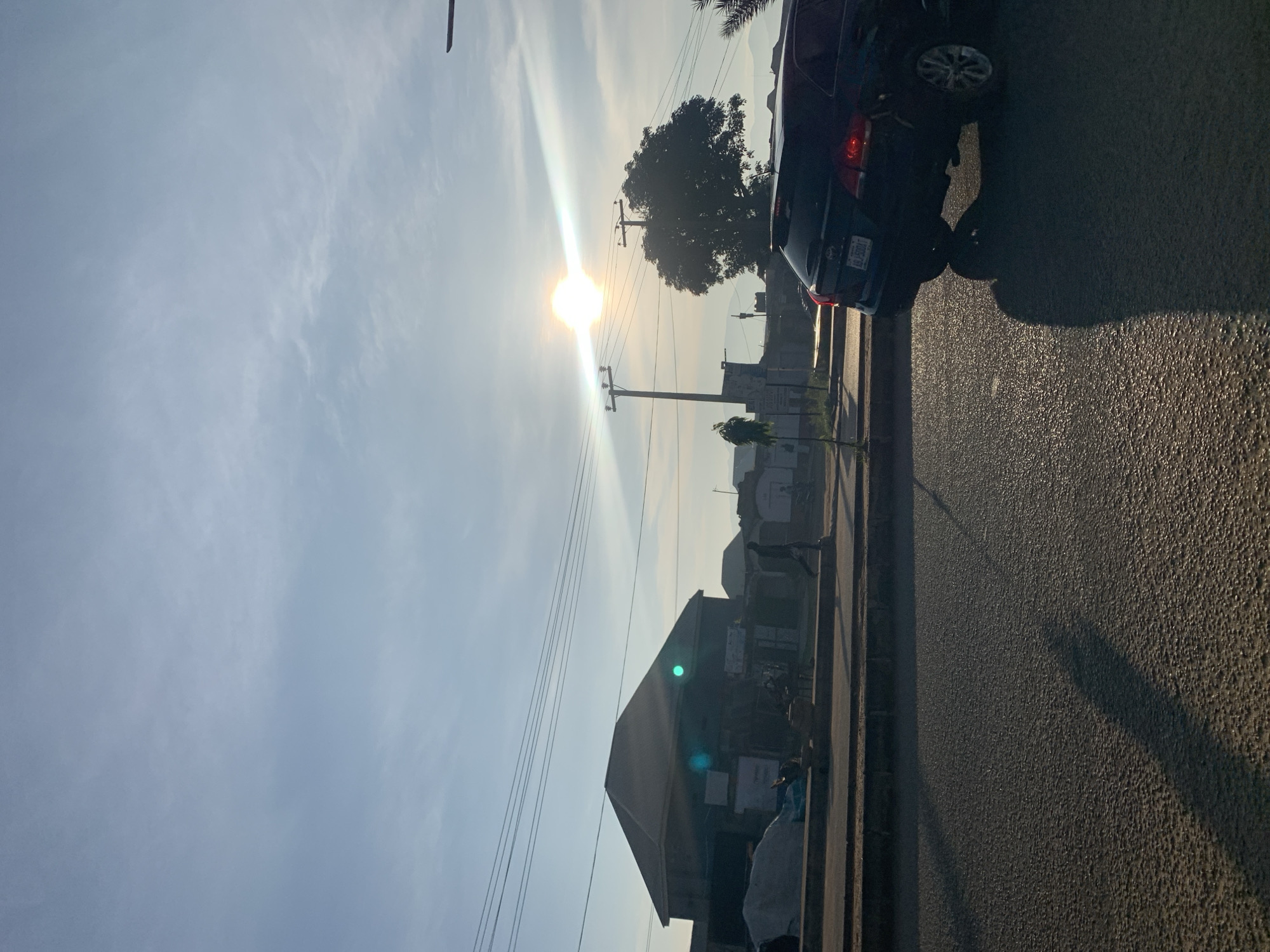 A low-angle, eye-level shot depicts a street scene at dusk in Rafin Par, Nigeria. The sun, positioned high in the sky, casts a bright glare, creating lens flare across the image and illuminating a dark-colored SUV in the foreground on the right. The SUV, with its taillights illuminated, is angled towards the viewer, casting a long shadow on the textured asphalt road. In the mid-ground, a sidewalk runs parallel to the road, with several buildings visible. One prominent building, a two-story structure with a gabled roof, is situated on the left. To the right of this building, a large tree with dense foliage stands tall. A lone pedestrian is walking on the sidewalk in front of the buildings. Utility poles with overhead wires are scattered throughout the scene, indicating infrastructure. The sky above is a pale blue, with wispy clouds and the intense brightness of the sun dominating the upper portion of the frame. The overall impression is a view of a street in a Nigerian city as daylight fades.
