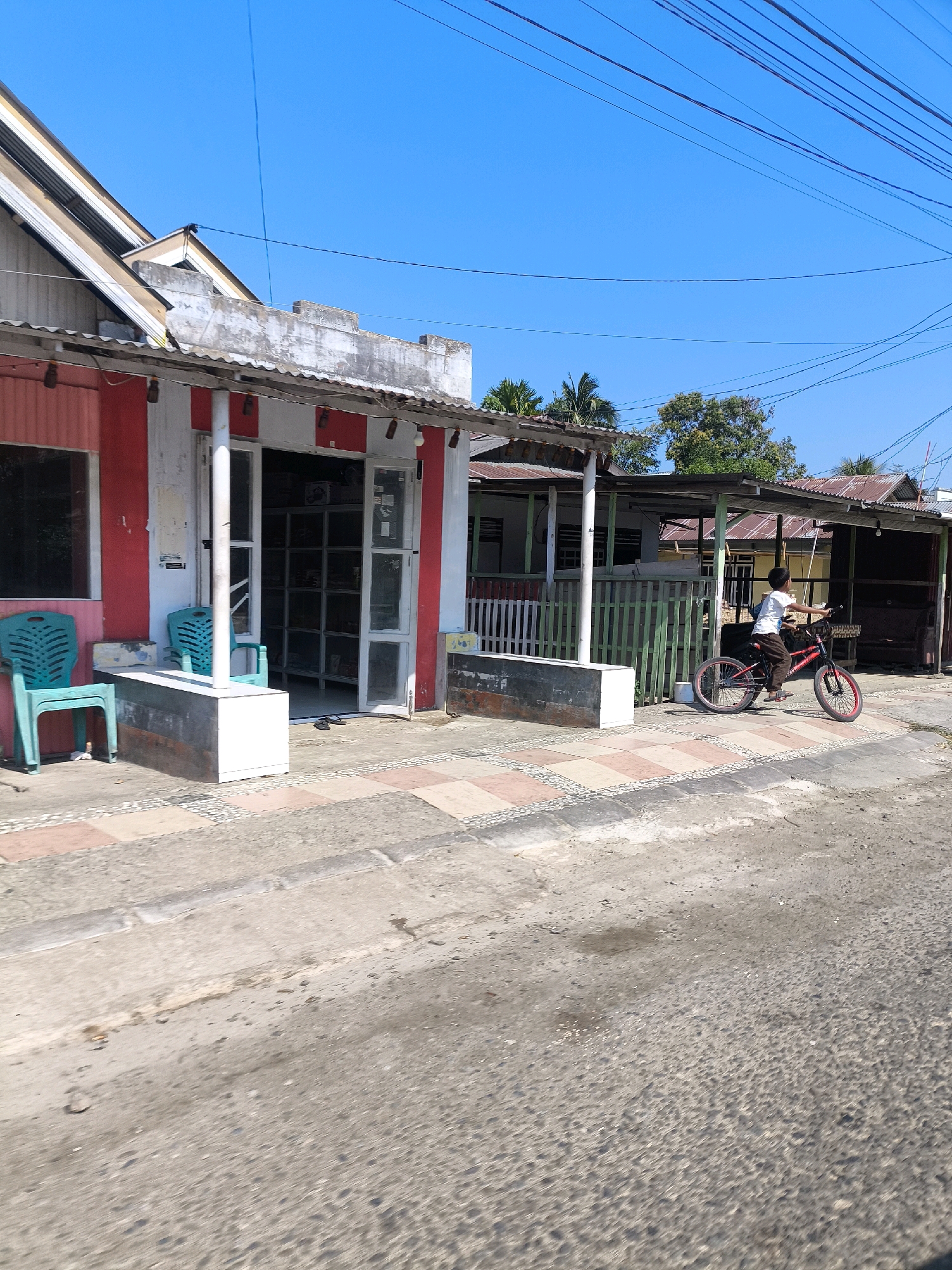 The image depicts a sunny day scene on a street in Tilamuta, Indonesia. In the foreground, a dirt and paved road transitions to a sidewalk with a patterned tile border. A young boy is riding a red bicycle with black tires and red rims on the sidewalk. He is wearing a white t-shirt and brown pants. To the left of the boy, a small, red and white building with an open doorway and windows appears to be a shop. Two green plastic chairs are placed on the sidewalk in front of this building. Further down the street, more buildings with corrugated metal roofs are visible. The sky is clear and blue.