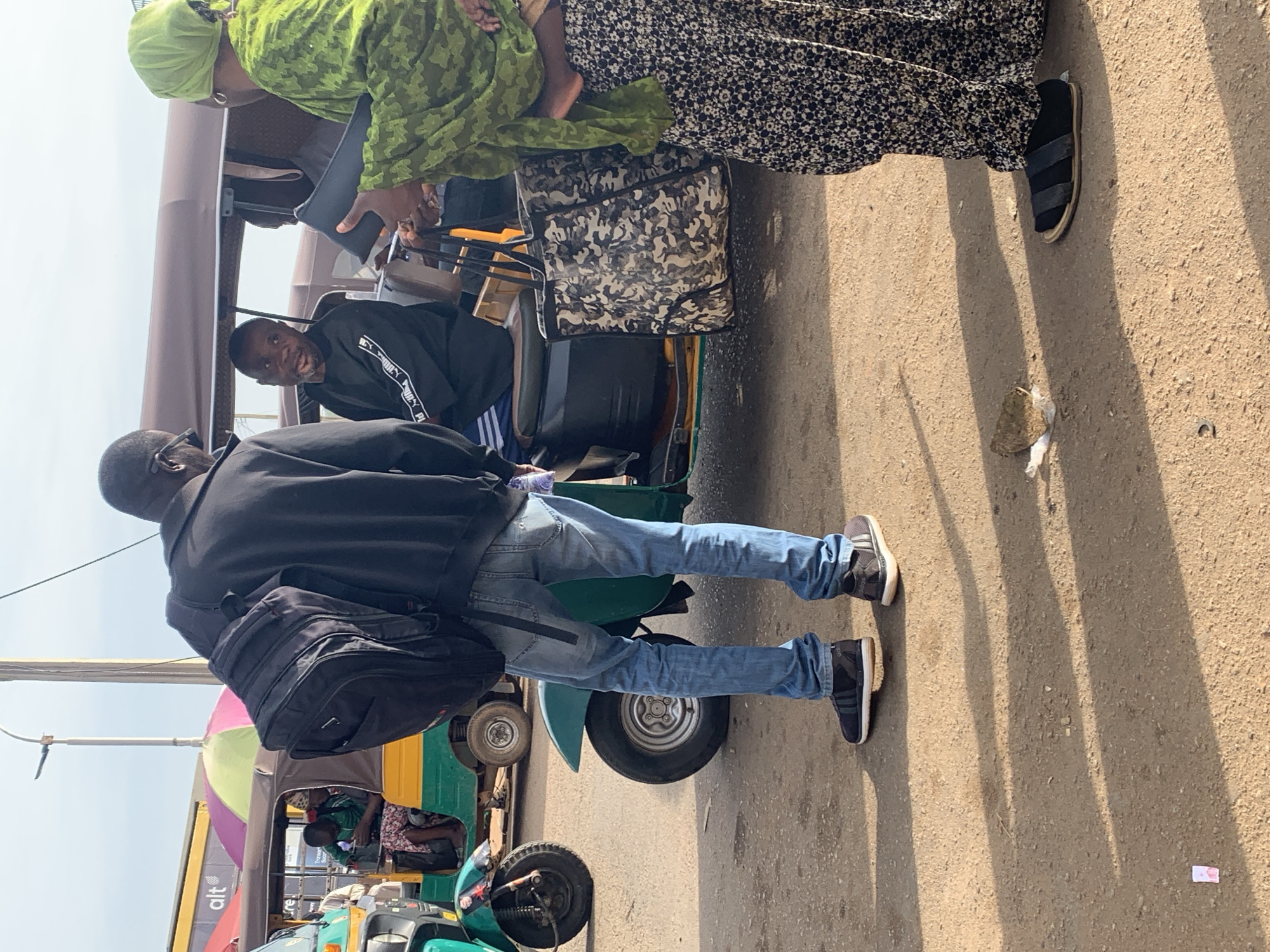In Gura Top, Nigeria, a group of people are gathered near several auto-rickshaws on a dirt road. A man wearing a black jacket and blue jeans stands with a black backpack slung over his shoulder, facing a group of people in one of the rickshaws. He appears to be speaking with the occupants. Inside the rickshaw, a man in a black sweatshirt and another individual in a green head covering are visible. A woman on the right side of the frame, wearing a green headscarf and a long patterned skirt, holds a black clutch bag. The scene suggests a moment of social interaction or transaction in a busy public space, possibly a transportation hub or marketplace. The rickshaws, typical of the region, are parked closely together. The ground is unpaved and dusty, indicative of a rural or semi-urban environment. The lighting suggests it is daytime, with sunlight casting shadows on the ground.