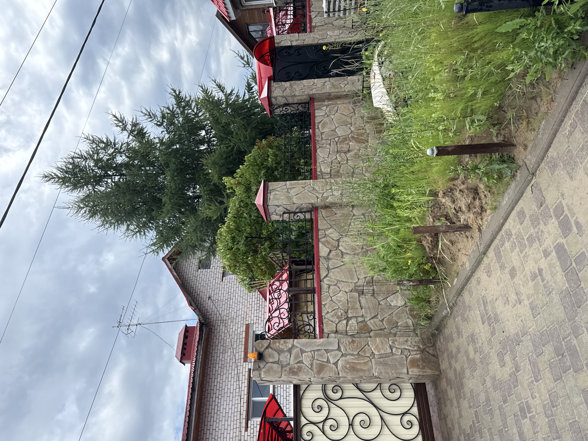 The image captures a section of a residential property in Kazan, Russia, on a cloudy day. The foreground shows a paved driveway made of interlocking rectangular stones in shades of gray and beige. To the left, a decorative metal gate with swirling patterns and a cream-colored panel is visible. Adjacent to the gate is a stone pillar that is part of a larger stone wall.
The stone wall, constructed from irregularly shaped and sized light-colored stones, extends across the midground. It is topped with a black metal fence featuring intricate scrollwork. Red awnings protrude from the tops of some fence sections and from a building.
Behind the wall and fence, a substantial evergreen tree dominates the center of the frame, its branches reaching high into the sky. To the left of the tree, a portion of a light-colored brick house with a red roof and a red chimney with a decorative top is visible. To the right, a wooden-clad house with a red roof is partially visible.
The environment is characterized by overgrown green grass and weeds growing along the base of the wall and in the foreground. Several small metal posts are embedded in the ground near the wall. Overhead, power lines stretch across the sky, and the sky is filled with dense, gray clouds. The overall impression is a semi-urban residential setting.