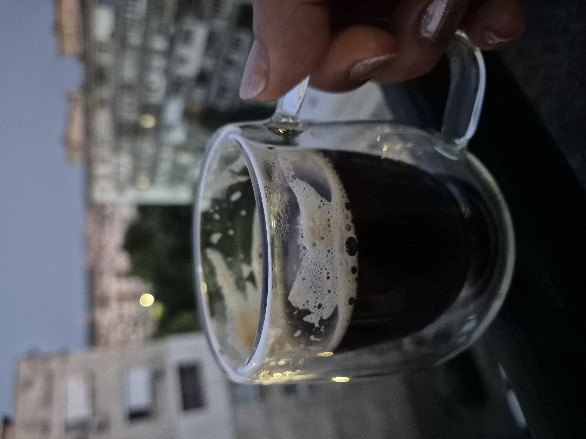 A close-up, eye-level shot shows a clear, double-walled glass mug filled with dark liquid and topped with a layer of foamy crema, likely espresso, positioned outdoors on a railing. The mug has a glass handle. A hand with manicured nails is holding the handle of the mug. In the background, out of focus, are several multi-story buildings and trees under a dusky sky, suggesting an urban setting during twilight. The lighting is dim, with some reflections visible on the mug.