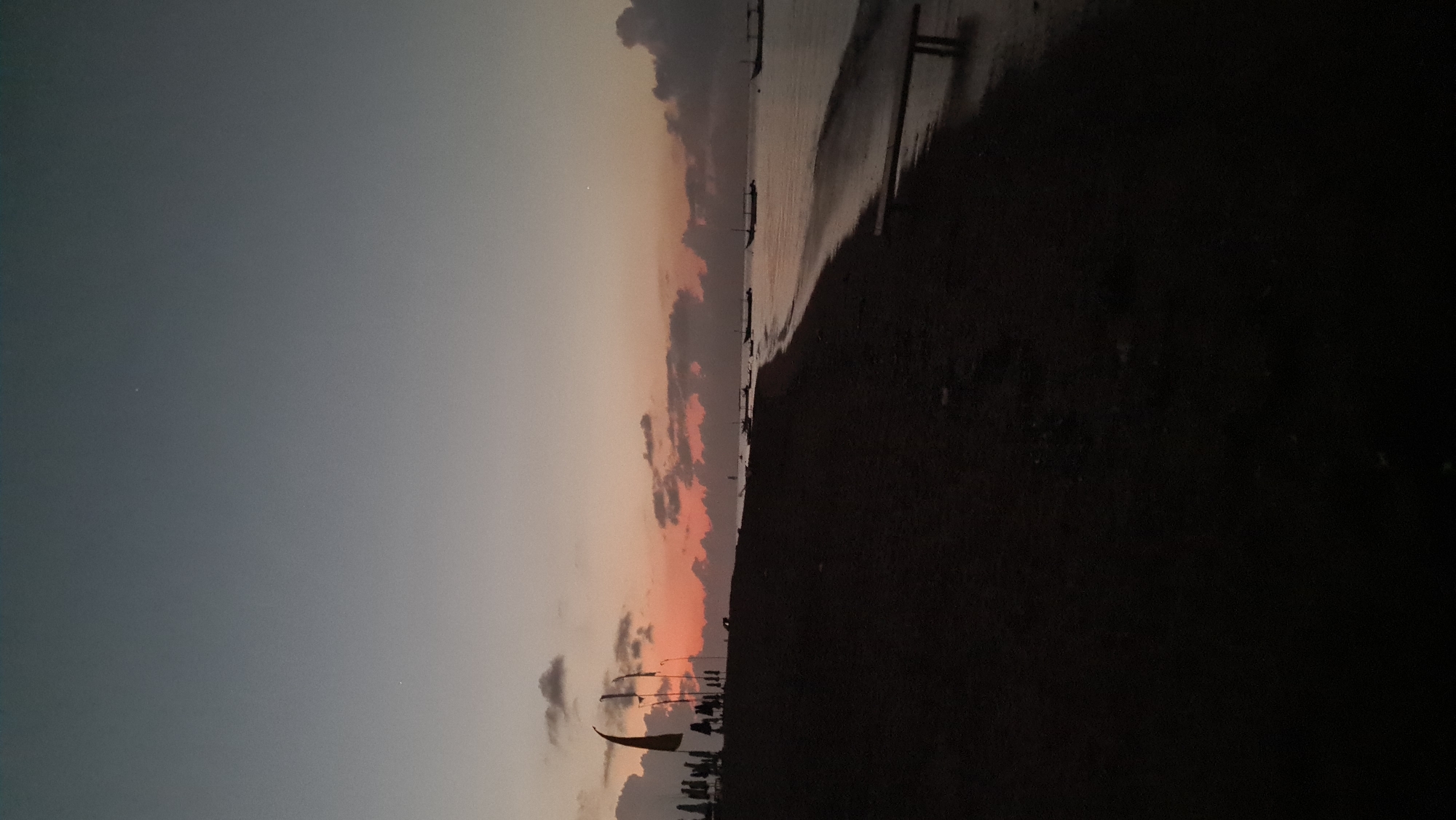 This image depicts a serene beach scene at twilight in Denpasar, Indonesia. The sky transitions from a deep dusky blue at the top to a soft, warm orange hue near the horizon, indicative of either sunrise or sunset. Wispy clouds are scattered across the sky, some catching the fading light.
In the foreground, a dark, sandy beach stretches along the coastline. Several figures, silhouetted against the brighter sky, are visible standing on the beach near the left side of the frame. Among them, what appear to be flags or banners are also silhouetted. Further down the beach, a simple wooden bench or structure is faintly visible near the water's edge.
The calm sea reflects the colors of the sky, creating a shimmering surface. Several small, traditional outrigger boats are anchored or drifting gently on the water, their silhouettes also dark against the lighter sea. The overall atmosphere is tranquil and peaceful, suggesting a quiet moment before dawn or after dusk.