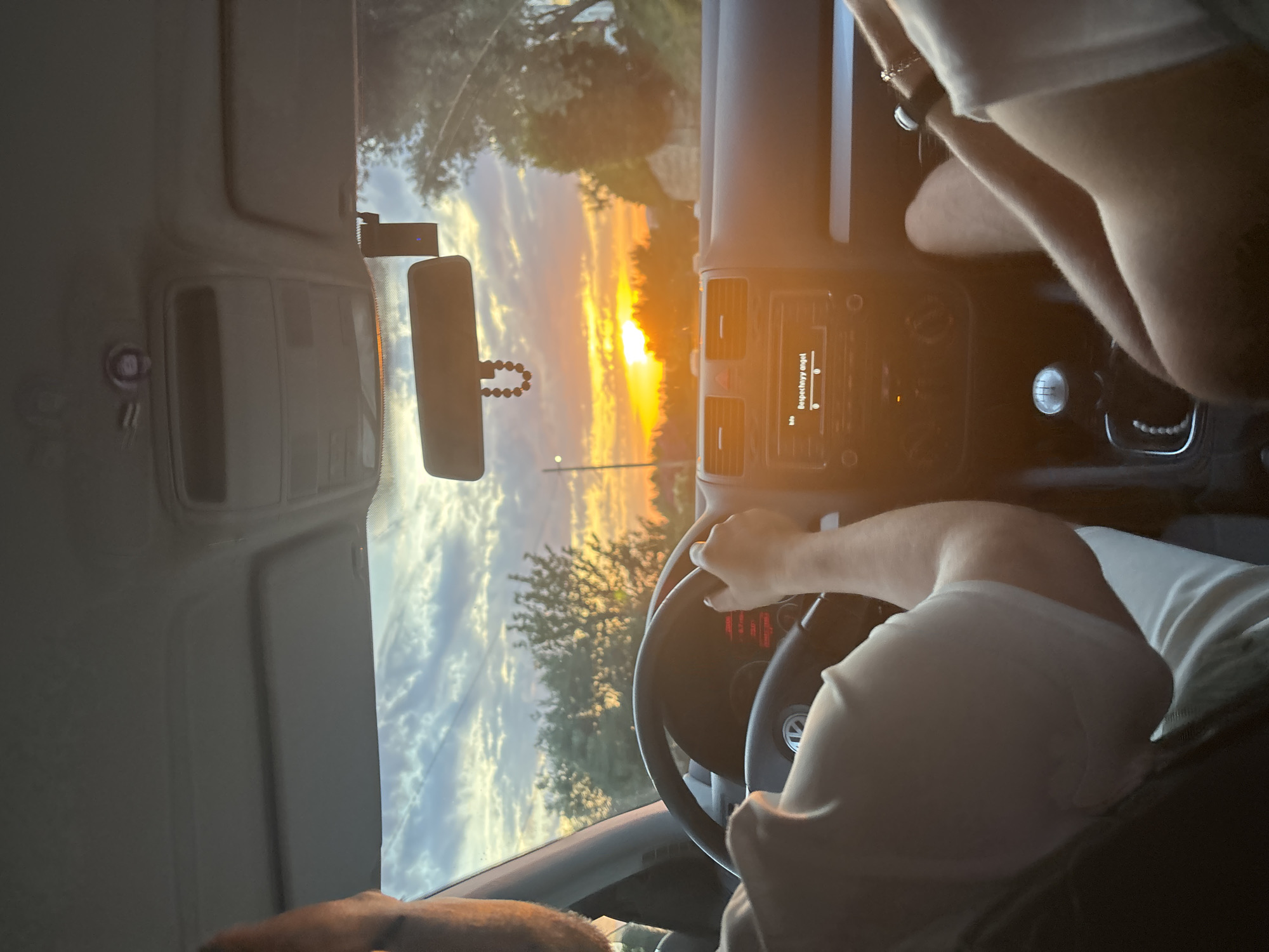 An interior shot of a vehicle, likely a Volkswagen due to the steering wheel emblem, captures a sunset scene in Gornji Katun, Montenegro. The driver, an adult male wearing a light-colored short-sleeved shirt, is positioned to the left, with his right hand on the steering wheel. A passenger, also an adult wearing a similar light-colored shirt and a wrist-worn accessory, is visible on the right, with their lower body and right arm in frame.
The car's interior features a dark gray dashboard, a manual gear shift with a 5-speed pattern, and an infotainment system displaying "Info" and "Bespechnyy angel" (Carefree angel). A rearview mirror, adorned with a beaded rosary-like object, hangs from the ceiling, which is light gray. A black dashcam or similar device with a blue indicator light is attached to the windshield to the right of the rearview mirror.
Outside the vehicle, a prominent golden-orange sun is setting on the horizon, casting a strong glare and illuminating the sky with vibrant orange, yellow, and light blue hues. Scattered clouds are visible against the colorful sky. Silhouetted trees line the road ahead, and a utility pole or thin vertical structure is discernible on the left side of the frame.