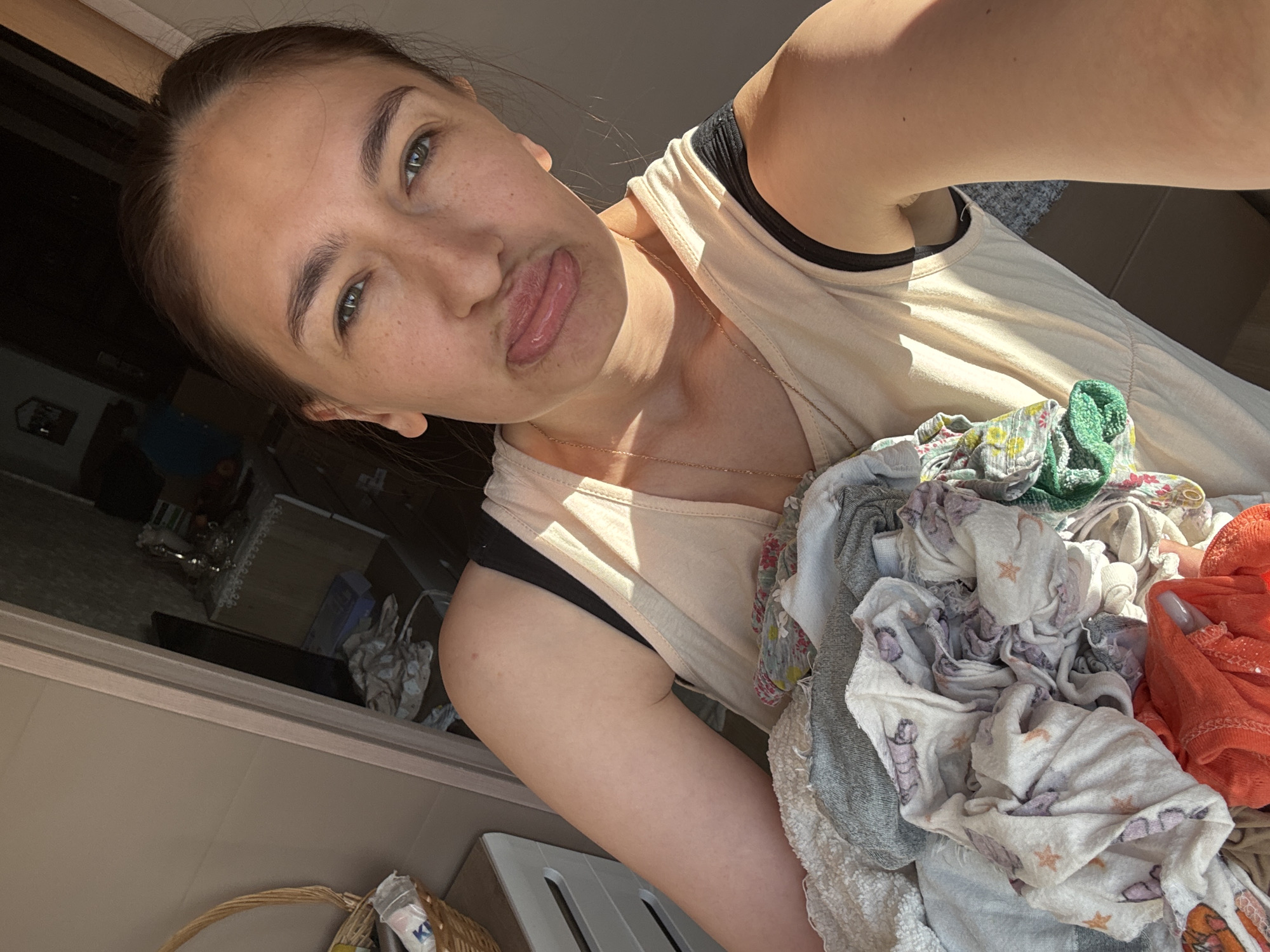 The image is a selfie taken by a woman indoors. She is holding a pile of laundry in her right arm, close to the camera. The laundry consists of various colored cloths, including gray, white with star and unicorn patterns, green, and orange. She is wearing a light-colored tank top over a black bra. The woman has fair skin, dark hair pulled back, and is sticking her tongue out slightly. Sunlight is illuminating part of her face and shoulder.
In the background, visible through a doorway or window, is a shelf with glass decorations. Below the doorway is a white chair and a wicker basket with objects inside. The location is indiscernible. There is no indication this image was taken in Sochi, Russia.