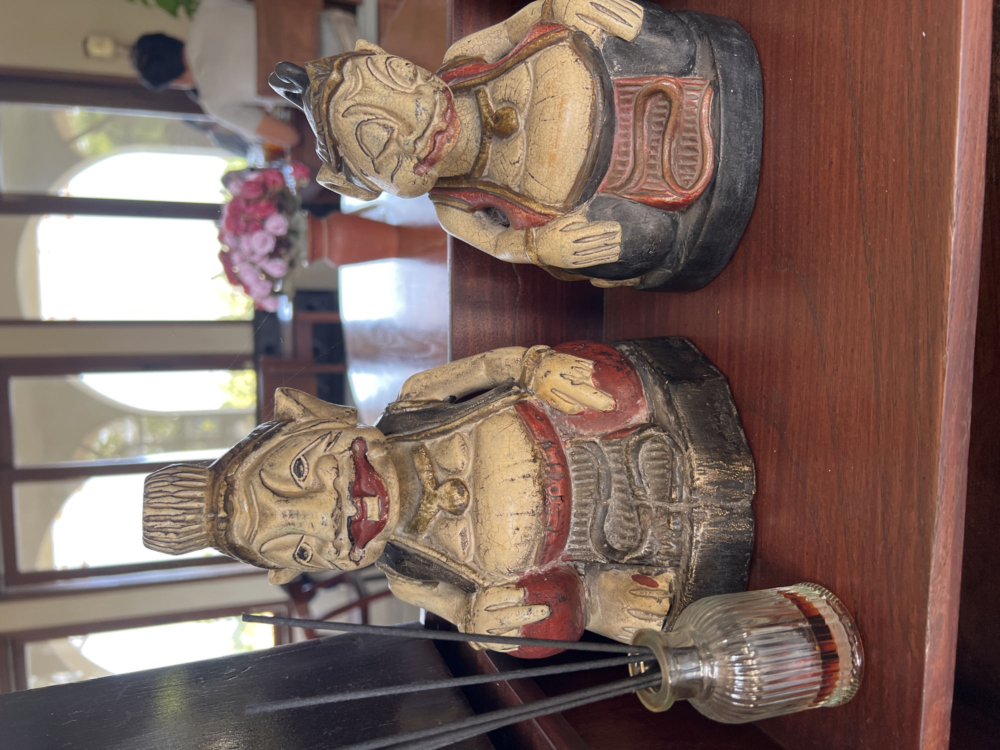 Two carved wooden figurines are placed on a wooden shelf. The figurines, likely of Balinese origin, depict figures with exaggerated features, including prominent mouths and bulbous stomachs. They are dressed in traditional attire with red sashes and gold ornamentation. To the left of the figurines, a small glass bottle filled with dark liquid and several thin black incense sticks is visible. In the background, a blurred scene suggests an indoor setting with large windows, possibly a restaurant or cafe, with tables, chairs, and a vase of pink flowers. A person is visible in the background, seated at a table. The overall impression is of a display of decorative items in a public or hospitality space.