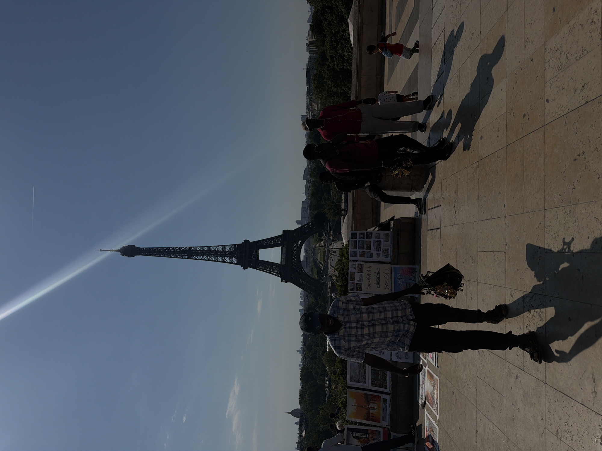 The image depicts the Eiffel Tower in Paris, France, under a bright, sunny sky with a prominent sun glare. In the foreground, several individuals are visible. A man in a plaid shirt and dark pants is walking towards the viewer, carrying a bag. Behind him, other people are standing or sitting, some appearing to be vendors with artwork displayed. Further in the background, a young child in a red shirt is playing, and another family is walking. The scene is set on a paved area with ornate stone railings. The city's skyline is visible in the distance, with trees and other buildings forming a backdrop to the iconic tower.