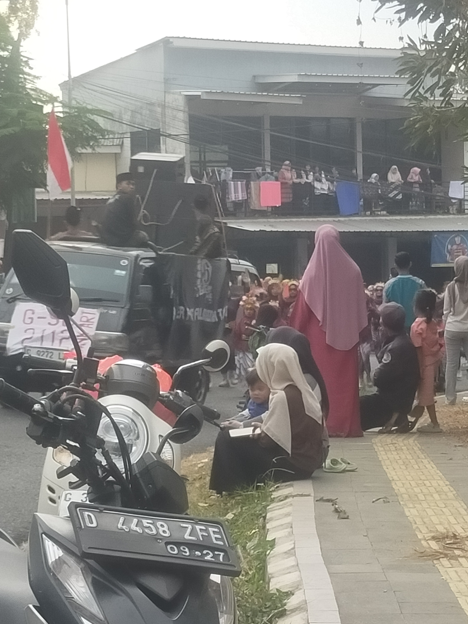 The image shows a street scene in the city of Tuwel, Indonesia. In the foreground, a motorcycle is parked, with its license plate reading "D 4458 ZFE 09-27". Behind the motorcycle, a black pickup truck is visible, with a large speaker system mounted on its roof and back. A black tarp with white text, possibly a banner, is draped over the back of the truck.
A group of people are gathered around the truck, some appearing to be participants in an event or gathering. Several individuals are wearing headscarves, indicating a predominantly Muslim population. In the background, a building with multiple stories and balconies is seen, with people visible on the upper floors. A red and white Indonesian flag is flying on a flagpole to the left of the truck. The sidewalk is paved with gray tiles and bordered by grass and a low wall. The overall impression is of a public gathering or event taking place on a street.