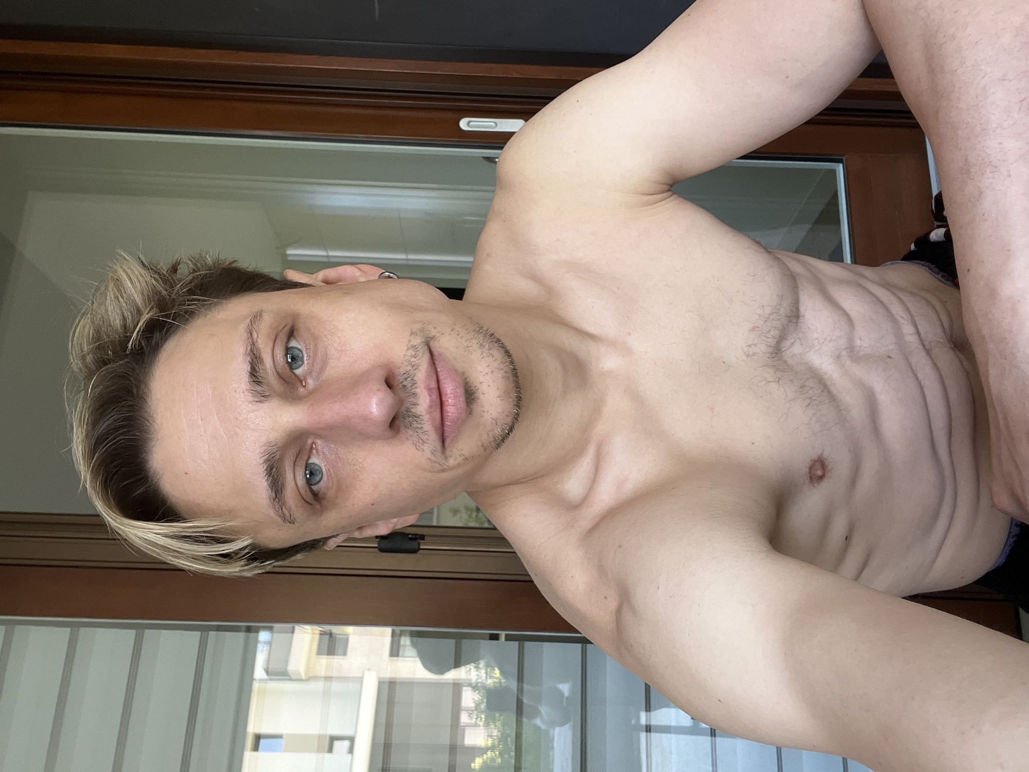 The media file shows a shirtless individual posing for a selfie, likely taken indoors near a window or balcony, as suggested by the reflection of a building and trees in the background. The individual has blonde hair, blue eyes, and a light beard stubble. Their body is lean with visible abdominal muscles. They are wearing dark underwear. The overall mood appears casual and relaxed. The lighting suggests it could be daytime, possibly morning or afternoon, with natural light filtering in. The presence of a modern building in the reflection hints at an urban or suburban setting. There is no visible text in the image.