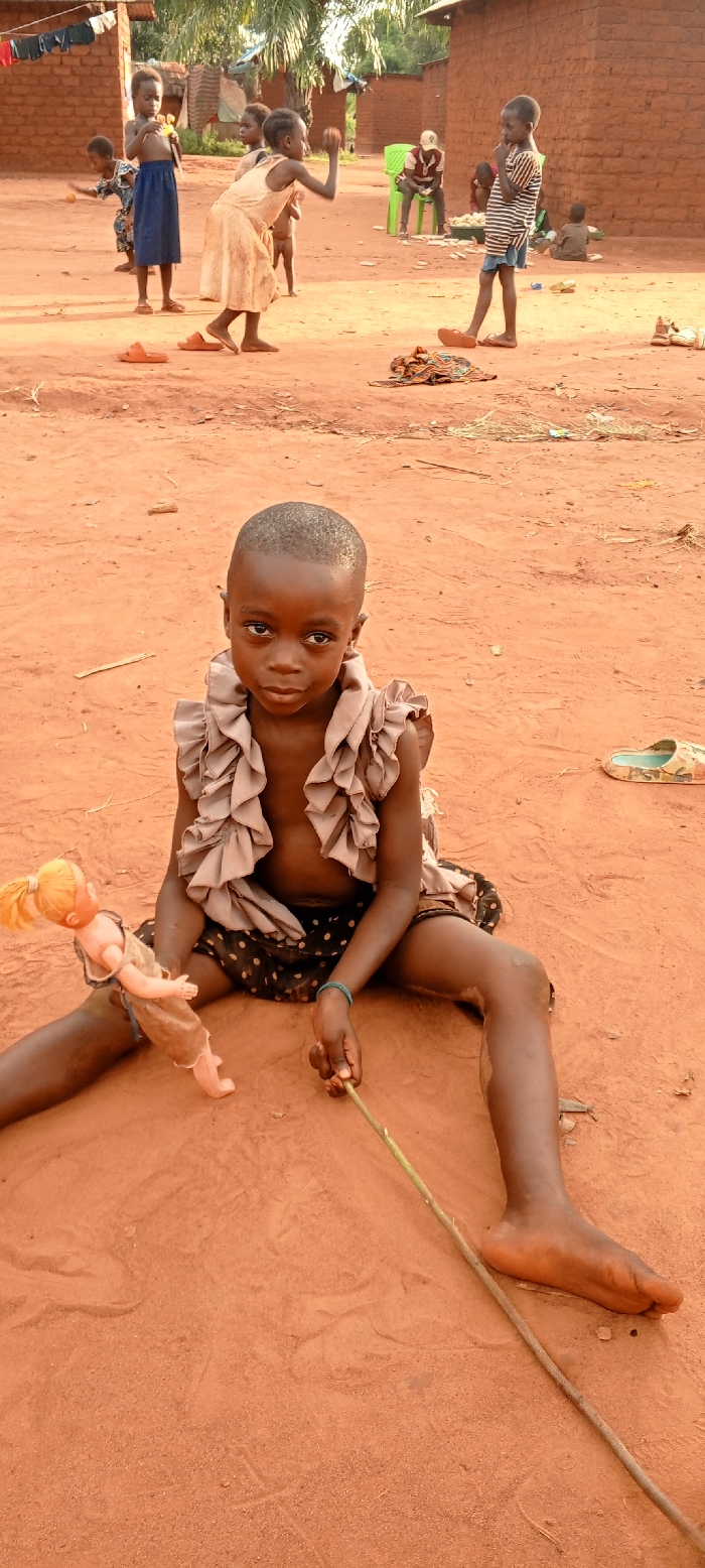 The scene depicts an outdoor setting in the city of Ngalula, Democratic Republic of the Congo, characterized by reddish-brown, dry earth as the dominant ground cover. In the foreground, a young child with dark skin and short dark hair sits directly facing the viewer, legs splayed on the ground. The child wears a light-brown, ruffled sleeveless top and a dark, polka-dotted skirt/wrap, and has a light-blue bracelet on their right wrist. The child's left hand cradles a blonde-haired doll, dressed in a light-colored garment, while the right hand holds a thin wooden stick resting on the ground.
In the mid-ground, several other children are visible. To the left, a child with a bare chest and a blue skirt stands, holding a yellow object. Center-left, another child in a light-colored dress is captured in motion, with their right arm raised as if throwing or catching. Further to the right, a child in a striped shirt and shorts stands observing. In the background, an individual wearing a red shirt and a white cap sits on two green plastic chairs. Additional figures are partially obscured by buildings or distance.
The background consists of several single-story structures constructed from unplastered reddish-brown bricks, topped with corrugated metal or thatched roofs. Clothes are hanging on lines near some of these buildings. Sparse tropical vegetation, including palm trees, is visible in the distant background. Various items are scattered on the ground, including orange sandals, a bundle of patterned fabric, a camouflage-patterned slipper, and small pieces of wood or debris.