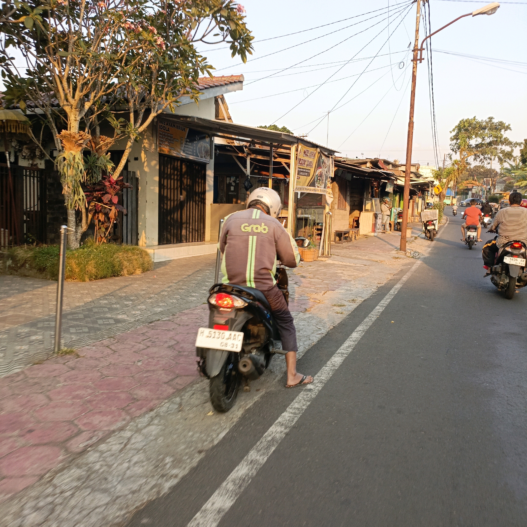 The image depicts a street scene in Salatiga, Indonesia, characterized by a row of roadside establishments on the left and a street on the right. In the foreground, a person wearing a white helmet and a gray jacket with green accents, featuring the "Grab" logo, is riding a black motorcycle. The motorcycle has a license plate with the inscription "H 5130 AAC 08-21". The person is wearing dark pants and flip-flops.
The roadside on the left features a sidewalk with red hexagonal tiles, bordered by grass and a metal pole. Behind the sidewalk are several small buildings with corrugated metal roofs and open fronts, displaying various signs, some of which appear to be menus for food establishments like "MIE AYAM KRIUK IDOLA". A tree with pink flowers is also prominent in this area.
The street on the right is paved and has a white line marking its edge. Other motorcycles are visible on the street, including one in the middle distance with a rider in a red shirt and another further ahead with a rider in a striped shirt. Overhead, a network of electrical wires crisscrosses the sky, supported by a rusty utility pole with a streetlamp. The lighting suggests it is late afternoon, with warm sunlight casting long shadows.