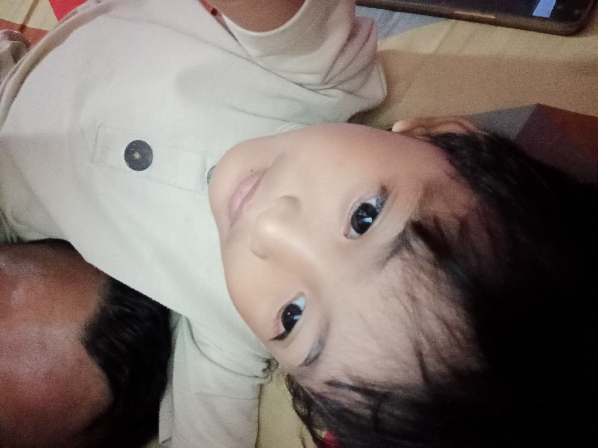 The image shows a close-up, tilted overhead view of a child's face and upper body, wearing a light-colored shirt. The child's head is angled towards the viewer, with their eyes open and looking directly at the camera. Part of another person's head and shoulder, with dark hair, is visible in the lower left corner, suggesting they are lying down next to the child. The background appears to be a bed or couch with a patterned surface. The overall lighting is dim. There is no discernible information about the location in Bloro, Indonesia within the image itself.
