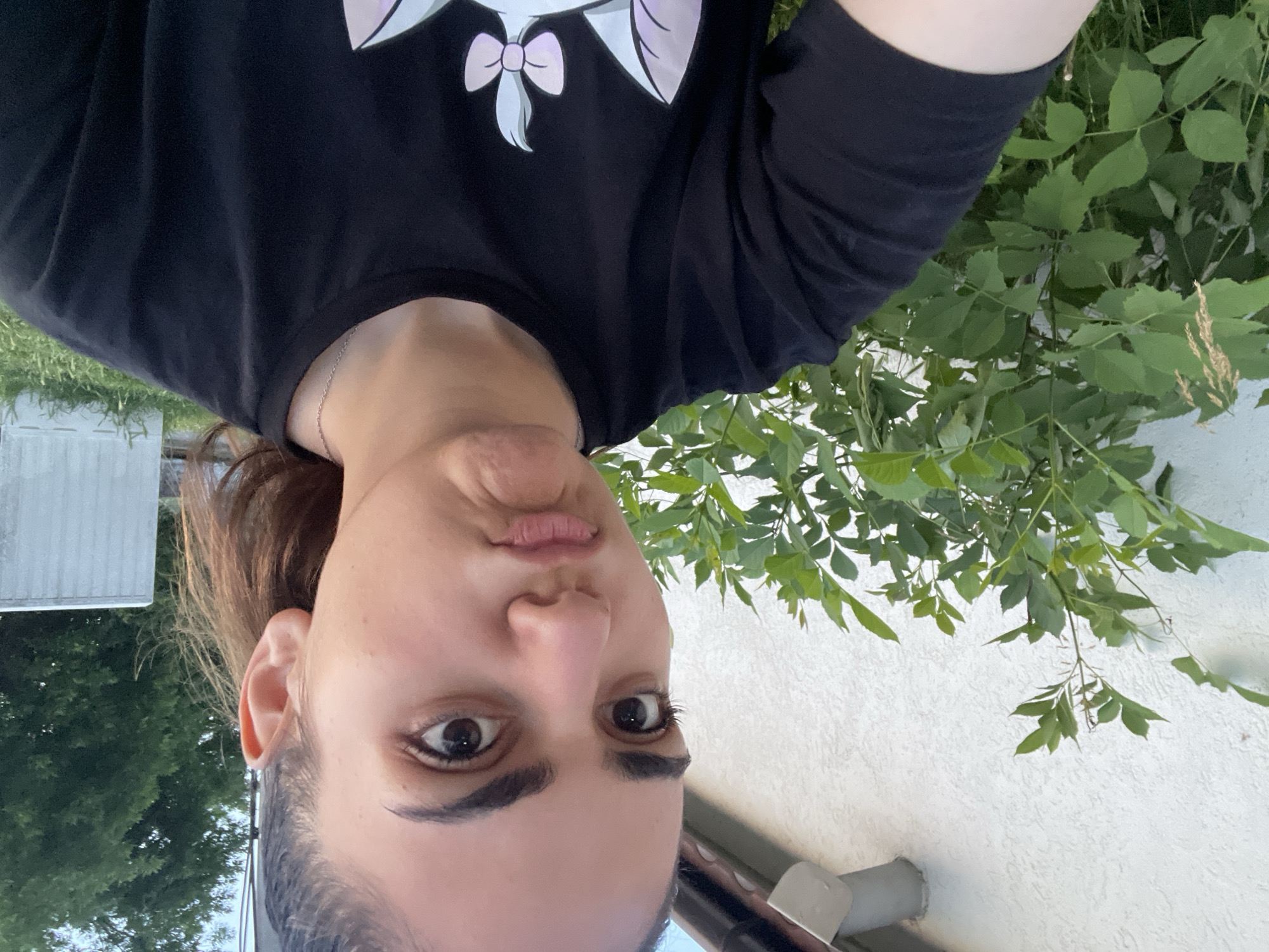 A young female is depicted in a close-up outdoor selfie in Debelt, Bulgaria. She faces the camera directly, displaying a pouting expression with pursed lips and a slightly scrunched chin. Her dark brown eyes are open wide, and her dark hair is pulled back, with strands visible near her right ear. She wears a black, short-sleeved t-shirt featuring a partial light-colored graphic design, possibly a feline character with a pink bow, visible on the lower left of the garment. A thin silver chain necklace is visible on her collarbone. The immediate background to her left consists of a textured, light-colored building wall. A grey drainpipe segment is visible near the top of this wall, below what appears to be a roofline. Dense green foliage, consistent with shrubs or bushes, occupies the mid-ground behind and to the left of the subject. In the far background to the right, dark green trees are visible alongside a white, corrugated panel structure situated above a patch of green grass. The scene is illuminated by natural daylight.