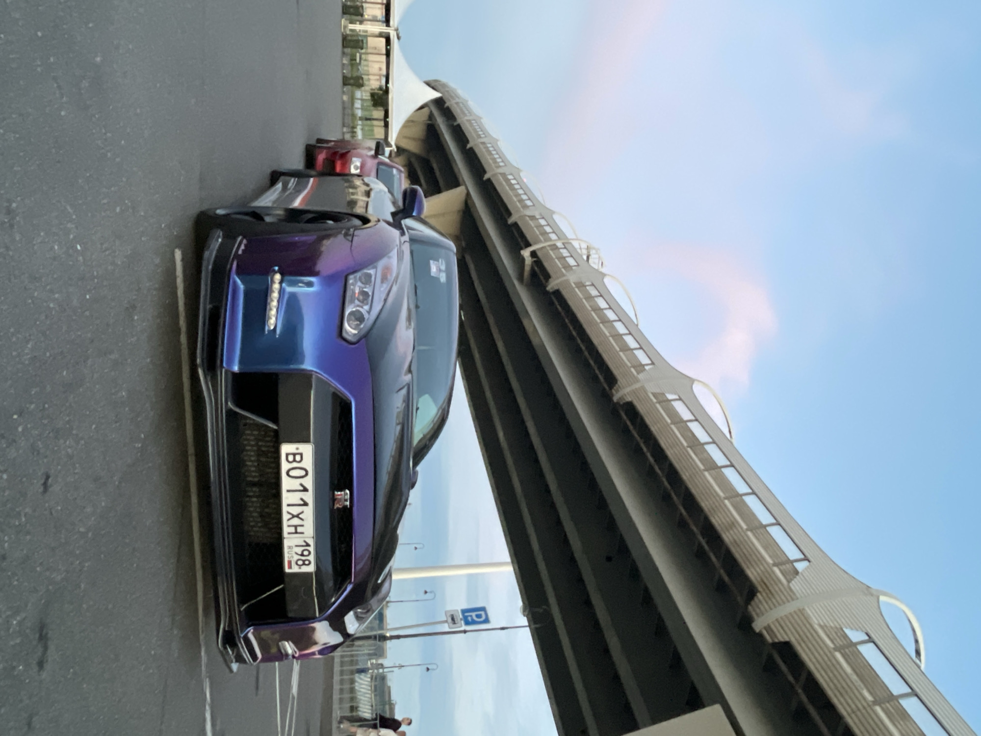A Nissan GT-R with a iridescent purple and blue paint job is parked on asphalt, with a red car behind it. The GT-R has a black hood and a license plate that reads "B011XH 198 RUS". A large, modern overpass or bridge structure dominates the background, with a clear blue sky above. In the far background, there are some white tents and a few people are visible. The lighting suggests it is either early morning or late afternoon.
