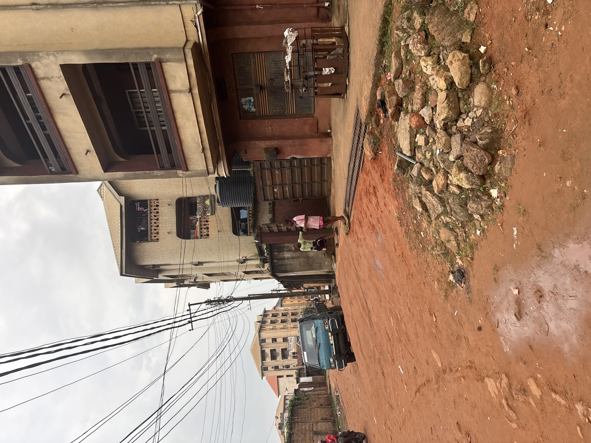 The image depicts a street scene in Onitsha, Nigeria, characterized by its reddish-brown, unpaved road. The environment appears somewhat neglected, with scattered rocks and debris on the roadside and in puddles of water on the ground. Several multi-story buildings line the street, some with balconies and exposed utility wires.
In the foreground, two children, a girl in pink shorts and a t-shirt and a boy in a green t-shirt and black shorts, stand on the side of the road. The girl appears to be pointing. A light blue truck is parked on the dirt road, facing away from the viewer. In the middle ground, more buildings are visible, along with a motorcycle with a rider dressed in red. In the background, additional buildings and more overhead wires are present. The sky is overcast and grey.