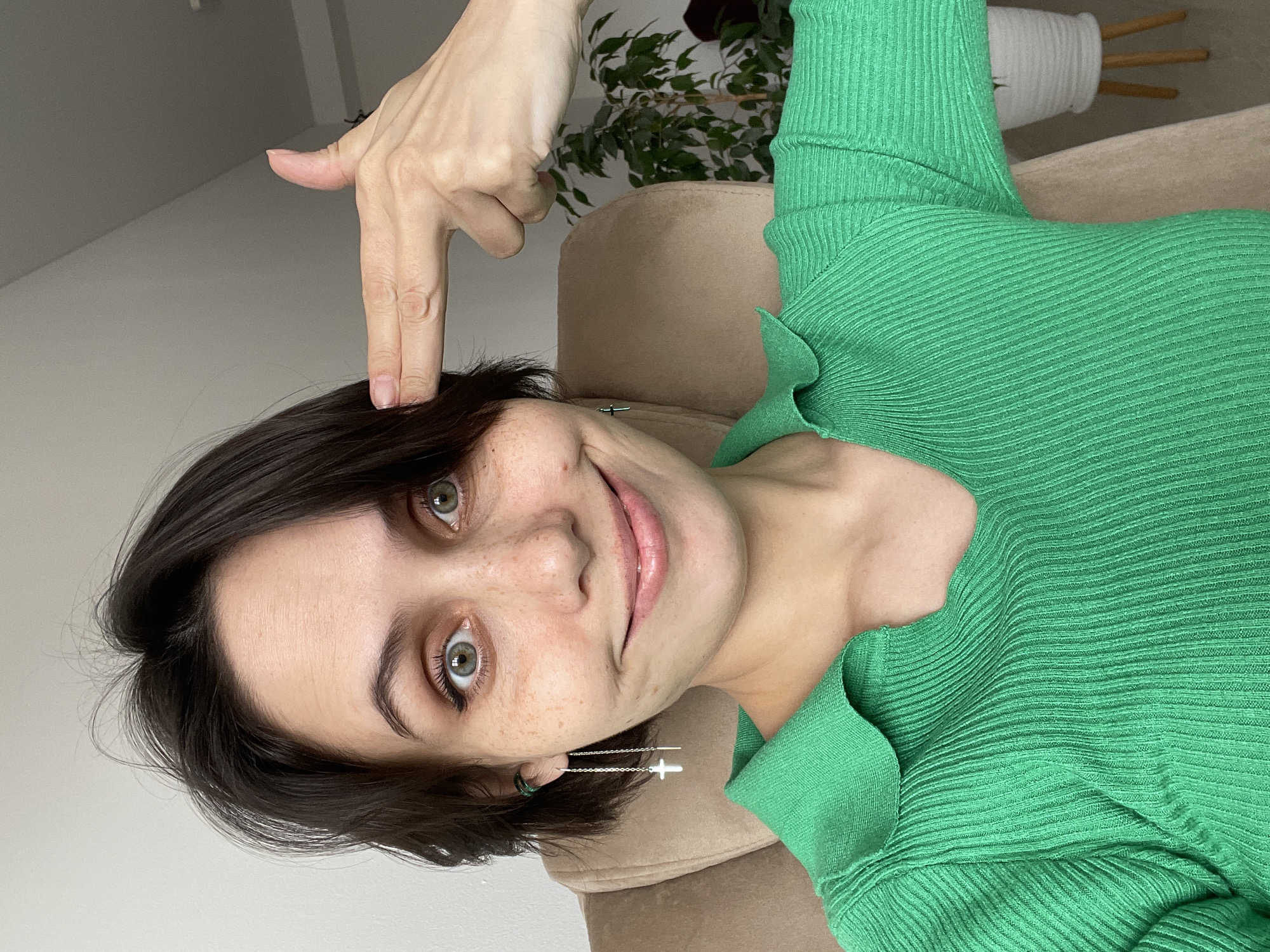 A young woman with short, dark brown hair and green eyes is prominently featured in a close-up, upward-angled shot. She has a light complexion with visible freckles across her nose and cheeks. Her lips are slightly parted in a soft smile, and she is wearing eye makeup, including eyeliner.
She is dressed in a ribbed, long-sleeved green top that appears to be made of a textured, possibly knit or woven, fabric. The top has a V-neckline with a ruffled or decorative edge. In her left ear, she wears a long, silver threader earring with a small cross design. Another small green detail is visible in her right earlobe.
Her right hand is raised, with her index finger and thumb extended, forming a "finger gun" shape, and pointed towards her right temple. Her fingernails are short and unpainted.
The woman appears to be lying down or reclining on a light brown, upholstered chair or sofa. The background is a minimalist indoor setting, with a plain light-colored wall visible. In the upper right portion of the image, a green potted plant with several leaves and a white textured planter can be seen, partially obscured by the woman's arm and clothing. The lighting suggests an indoor setting during the daytime. There is no visible text in the image.
The overall mood is playful, conveyed by the finger-gun gesture and her smiling expression. The location context provided is Izhevsk, Russia.