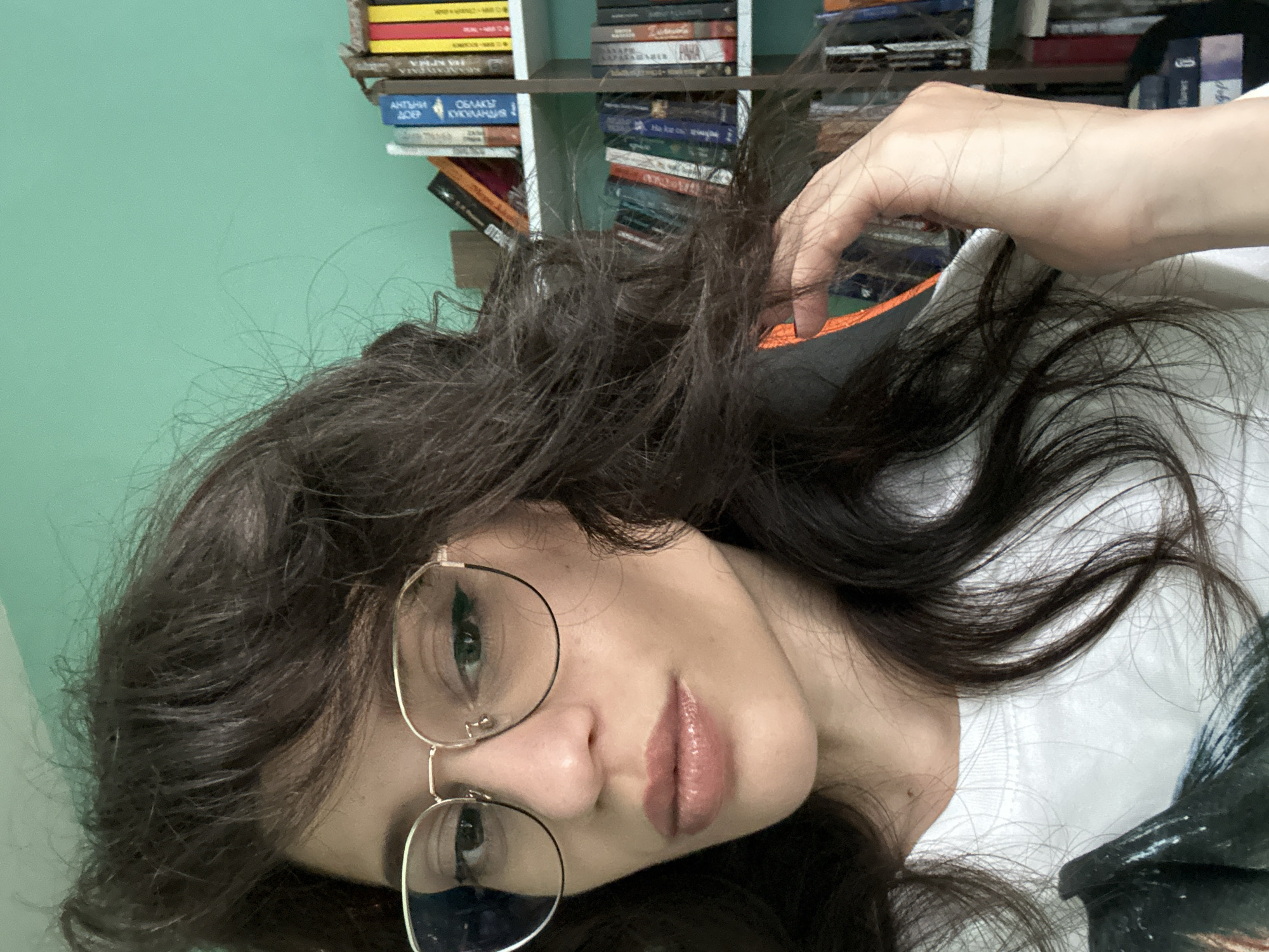 A medium close-up shot shows a woman with dark, wavy hair and glasses. She is wearing a white t-shirt. Behind her, a bookshelf filled with books is visible against a teal-colored wall. The image appears to be taken indoors.