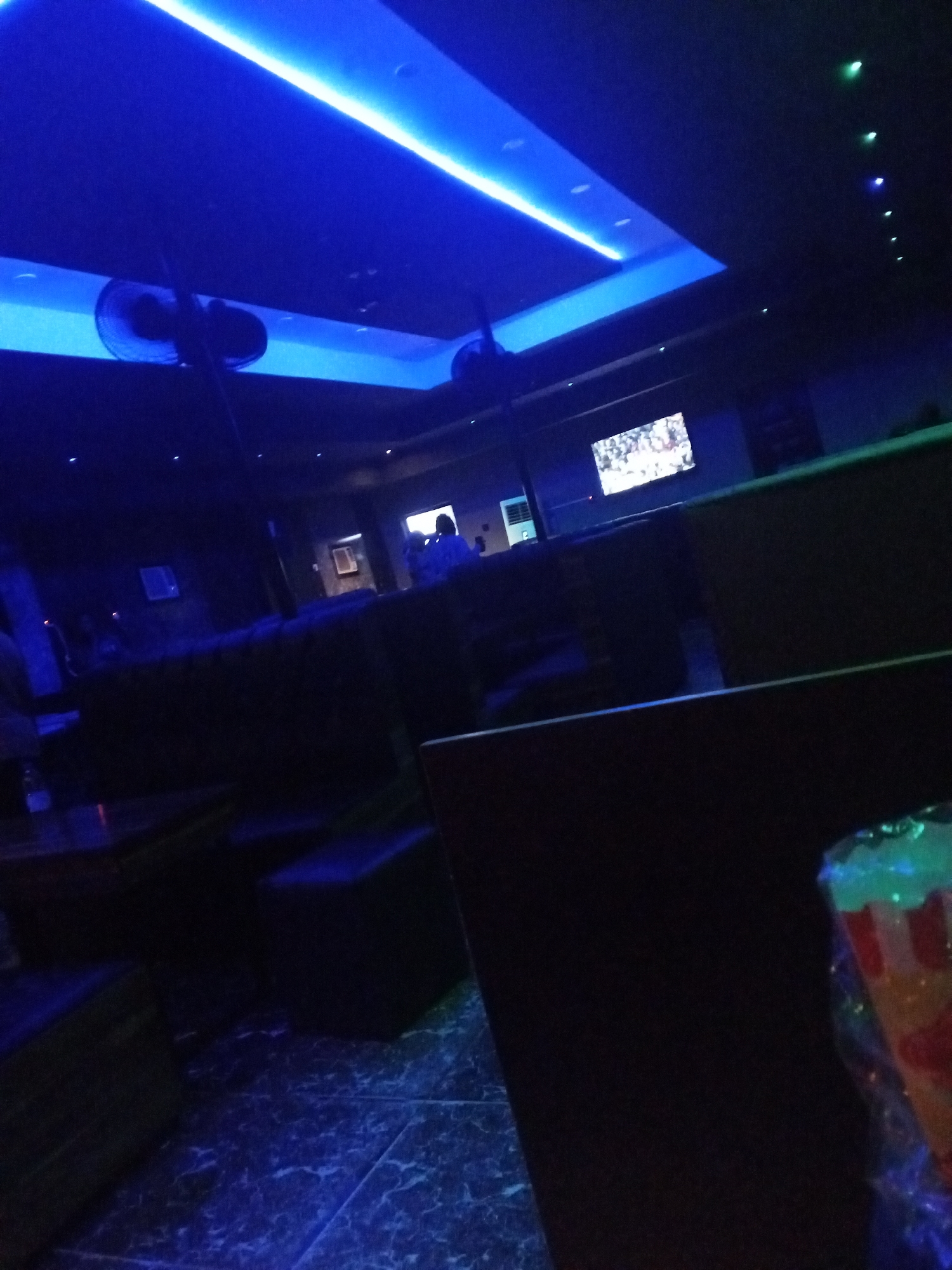The scene is an interior view of a dimly lit space, possibly a bar or lounge, located in Lokoja, Nigeria. The lighting is primarily blue, with a rectangular strip of bright blue light illuminating the ceiling. Two ceiling fans are visible above the couches in the darkness. There are several patrons in the background of the room, and a television screen is also visible. In the foreground, a dimly lit drink in a plastic cup sits on the right. A dark chair or table is immediately in front of the camera. The floor appears to be tiled.