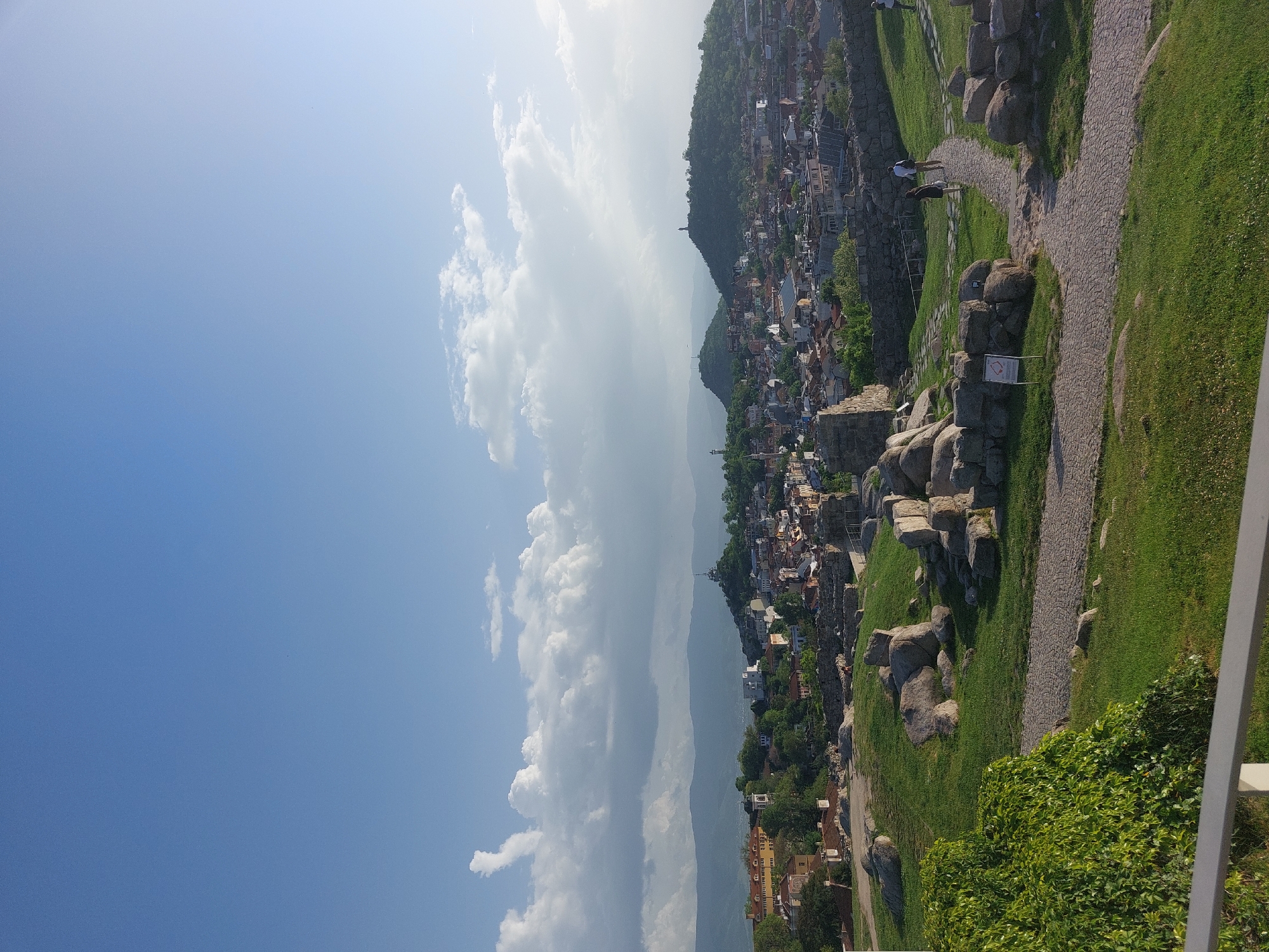 The image shows a view from the Nebet Tepe hill in Plovdiv, Bulgaria. A stone path winds through a grassy area near rock formations and ruins. A city is visible stretching along a mountain ridge, and there is a blue sky with cumulus clouds. Two people are walking on the path.