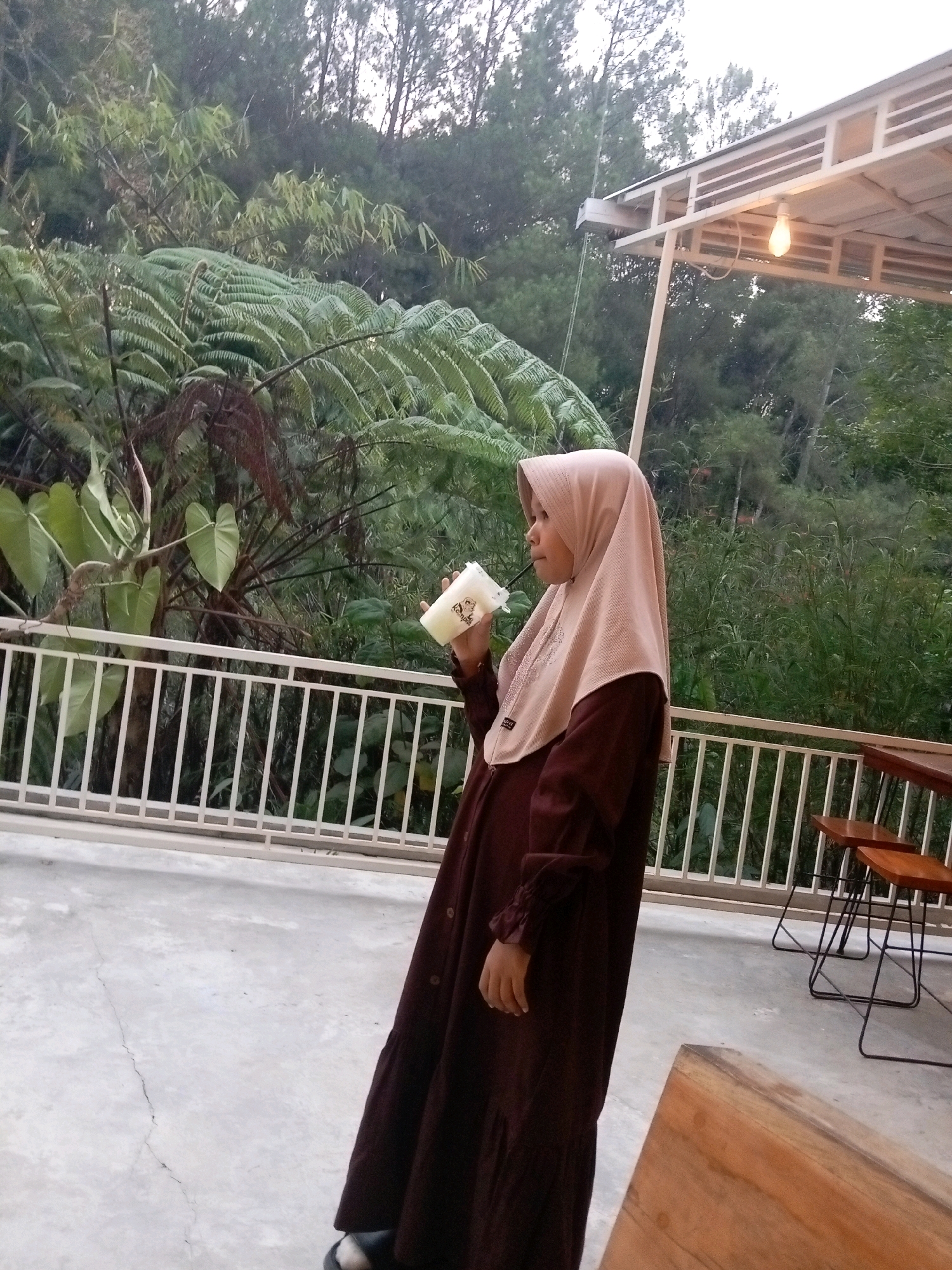 A young woman wearing a maroon dress and a beige hijab stands on a concrete patio, holding a white cup with a straw. She is facing left, looking away from the camera. Behind her is a white railing, lush green foliage including ferns, and a partially visible building with a light bulb hanging from its roof. In the background, trees can be seen. The setting appears to be an outdoor cafe or restaurant in a natural environment. The image was taken in Krisik, Indonesia.