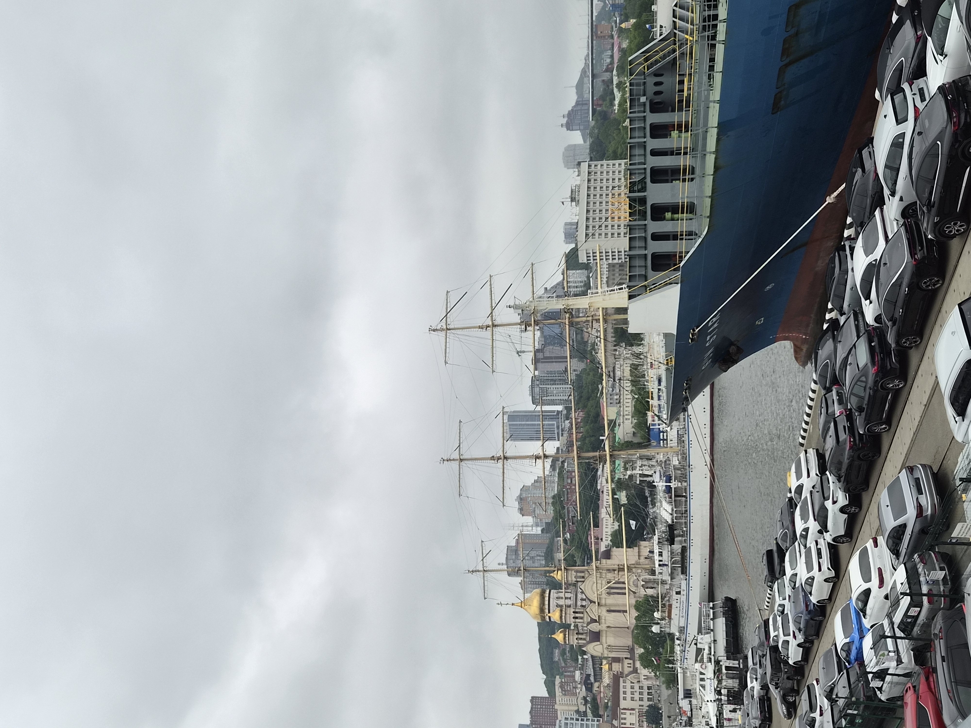 The image captures a port scene in Vladivostok, Russia, under an overcast sky. In the foreground, a large blue cargo ship named "SUN STAR" is docked, its hull showing a gradient of red and blue. The ship is secured by thick ropes. Adjacent to the ship, a large number of cars are parked in neat rows on the dock, indicating a car import or export operation. These vehicles are of various makes and models, predominantly in white, black, and silver.
In the middle ground, several other vessels are present in the water, including a tall ship with multiple masts. Behind the ships, the cityscape of Vladivostok unfolds. Notable structures include a prominent Orthodox church with golden domes, various multi-story buildings, and residential blocks climbing the hilly terrain. A suspension bridge is visible in the distance, a common landmark in Vladivostok. The overall atmosphere is that of a busy working port, with the city's distinctive architecture forming the backdrop. There are no visible people in the frame.