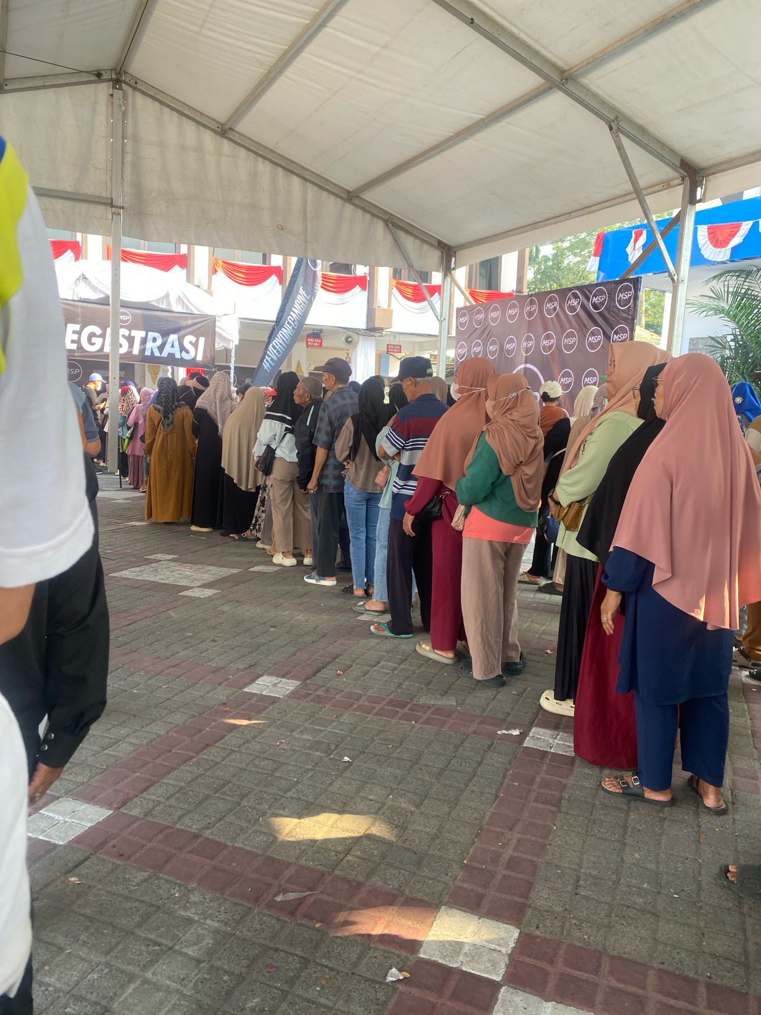 The image shows a line of people, mostly women wearing hijabs, standing under a tent. A banner in the background reads "EGISTRASI", suggesting a registration or check-in process. There are multiple smaller banners with the letters "MSP" in circles, likely representing a company or organization. The ground is paved with square tiles in a pattern of grey and reddish-brown. Red and white draped fabric is visible in the background, attached to buildings. The scene appears to be outdoors, possibly at an event or public gathering. The overall impression is one of orderly waiting.