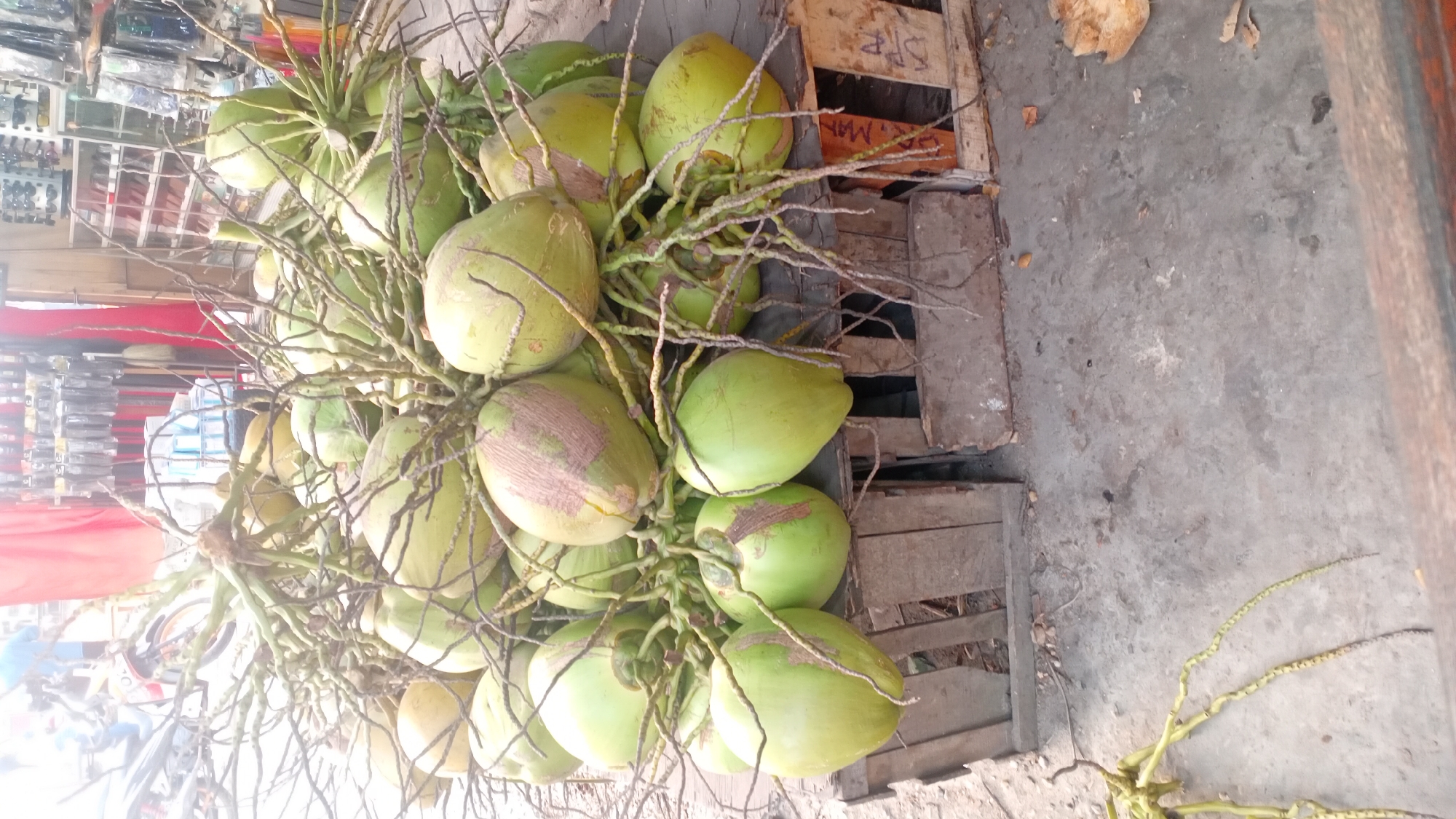 The image depicts a close-up view of a large bunch of green coconuts. The coconuts are attached to their stems and are piled on top of wooden crates. The background shows a market stall with various items displayed on shelves, including what appear to be packaged goods and eyewear. A red fabric hangs in the background, and a motorcycle is partially visible on the left side. The ground is concrete. This scene is likely from a market in Jambi, Indonesia, given the typical presentation of coconuts for sale in such environments.
