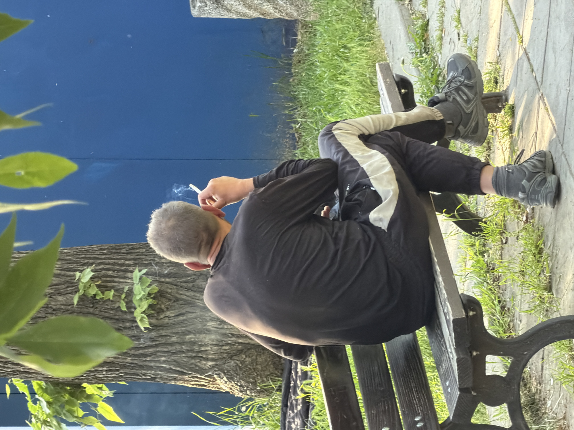 A man is sitting on a park bench smoking a cigarette. He is wearing a black tracksuit with a white stripe down the side and gray sneakers. The bench is in a park with grass and trees. The park is located in Sofia, Bulgaria.