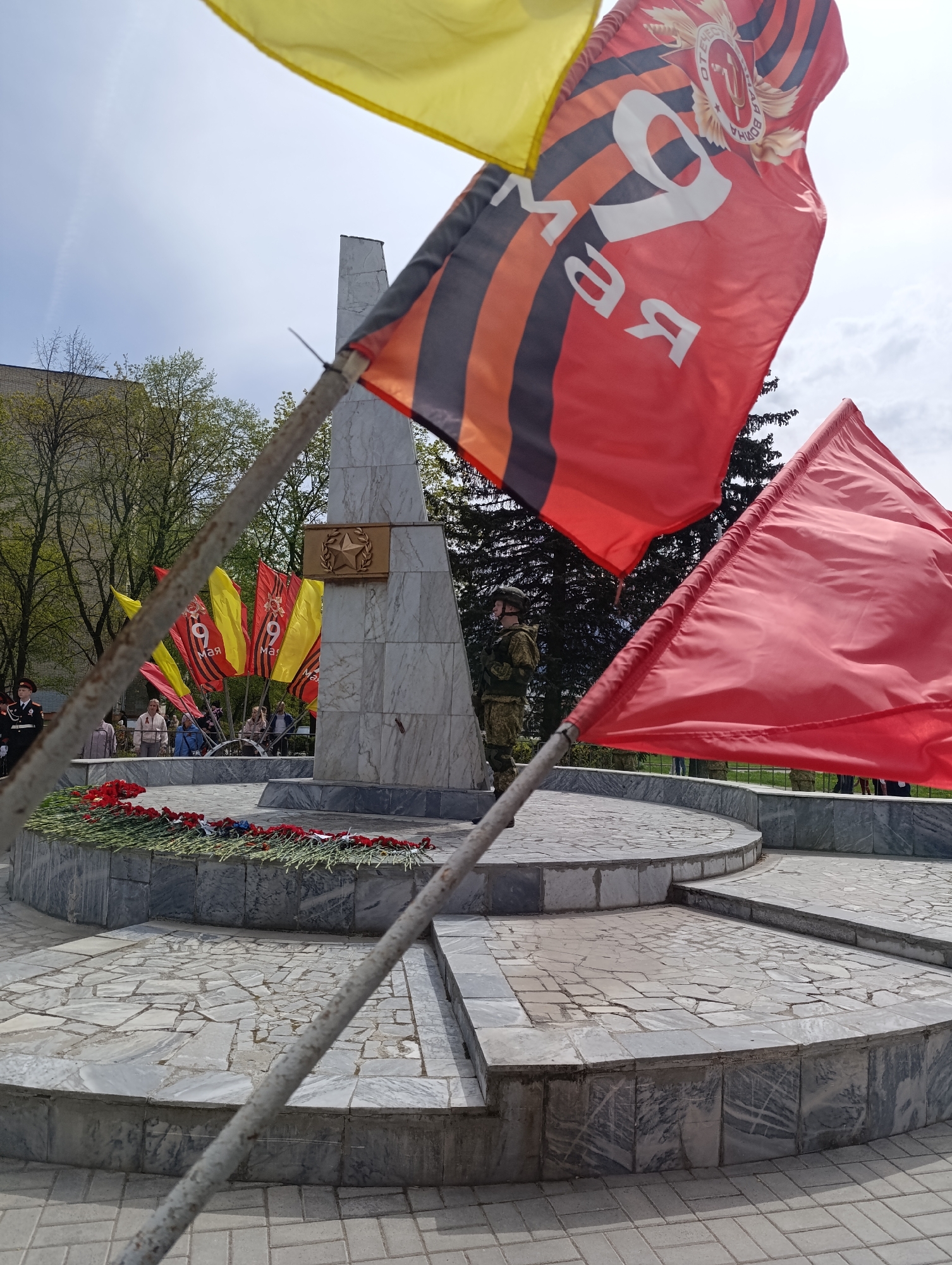 The image shows a memorial with a marble obelisk and a bronze plaque with a star. The memorial is surrounded by a multi-tiered circular platform made of grey marble tiles. Red and yellow flags are positioned in front of the memorial. A soldier in camouflage uniform stands guard near the obelisk. A wreath of red roses lies on the platform in front of the memorial. A group of people is visible in the background. The location appears to be outdoors, possibly a park or square in the city of Unknown, Unknown.