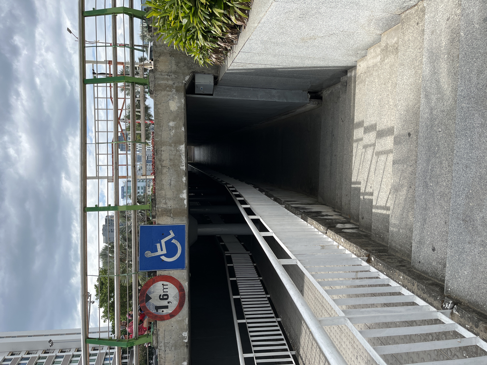 The image depicts the entrance to a pedestrian underpass or tunnel in an urban environment, likely in Đà Nẵng, Vietnam. The scene is dominated by concrete structures, including the tunnel entrance, walls, and descending stairs.
On the left side of the entrance, a long, white metal grated ramp or walkway descends into the dark tunnel, offering an accessible route. Parallel to this, on the right, are wide, textured concrete stairs also leading downwards. The tunnel itself is dark, suggesting artificial lighting is needed inside or it extends deeply underground.
Above the entrance, on a concrete pillar to the left, are two prominent signs. A blue square sign with a white wheelchair symbol indicates accessibility for people with disabilities. Below it is a red circular sign with a white background and black text "1,6m" (1.6 meters) with a diagonal line through it, signifying a height restriction for entry.
Further to the left, a green and silver metal barrier or railing structure separates the underpass area from the street level above. Through this barrier, a cloudy sky, distant multi-story buildings, and palm trees are visible. Small figures of people can be seen through the railings and near the signs. Lush green foliage is present along the upper concrete wall to the right.
The time of day appears to be daytime, with soft light from an overcast or partly cloudy sky casting shadows from the railings onto the stairs and ground, particularly on the right side. The concrete surfaces show some weathering and grime, indicating regular use.