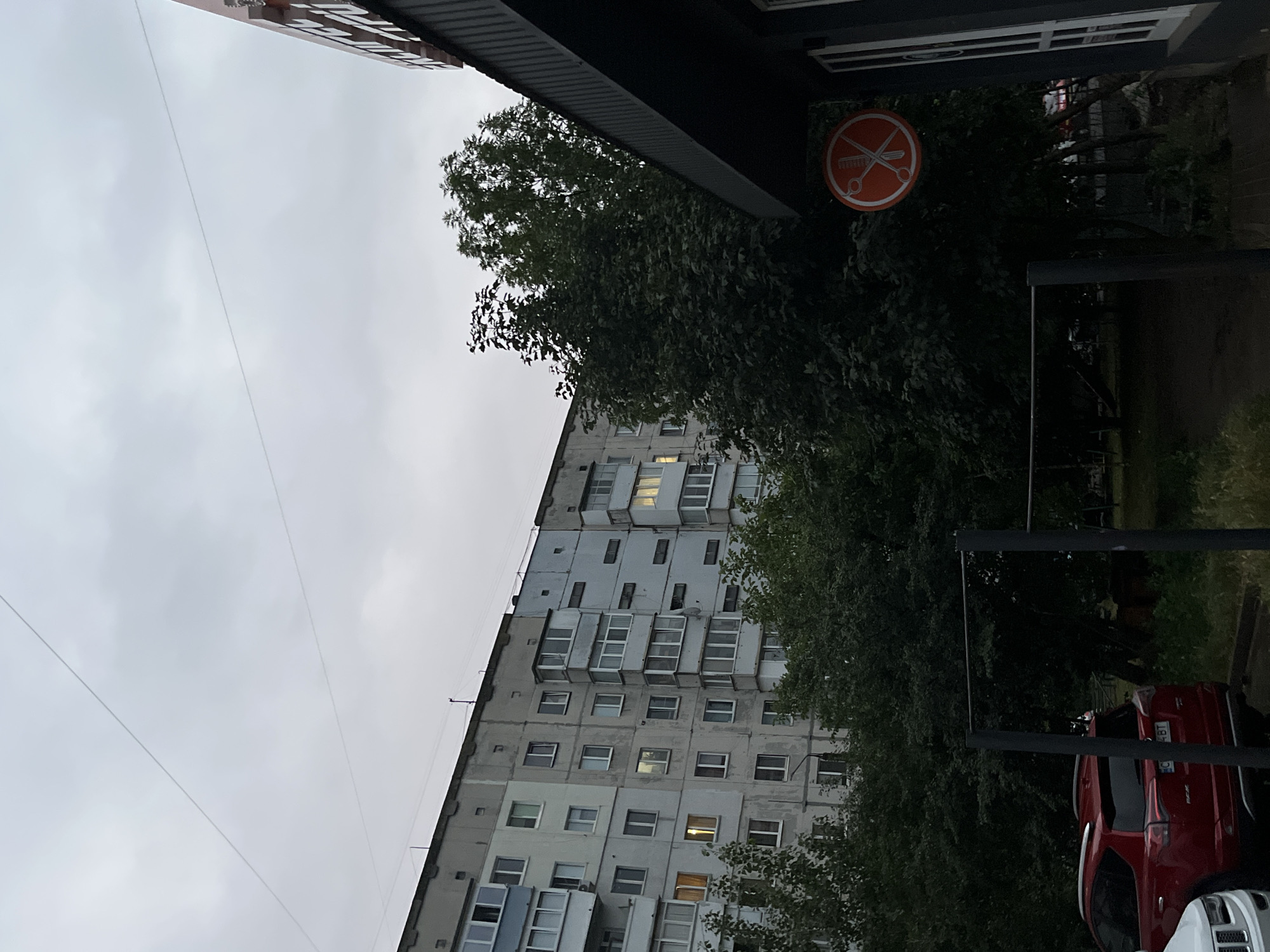 The image depicts an overcast sky above a multi-story apartment building and a commercial building with a visible sign. The apartment building is light-colored with numerous windows, some illuminated, suggesting interior lighting. Trees with dense foliage are situated in front of the building and to the right. To the right, a dark-colored commercial structure with a dark roof overhang is visible. Attached to the overhang is a circular orange sign featuring a white silhouette of scissors crossed with a comb, indicating a barbershop or hair salon. In the foreground, a red SUV is parked. The overall scene appears to be an urban setting in Chernivtsi, Ukraine.
