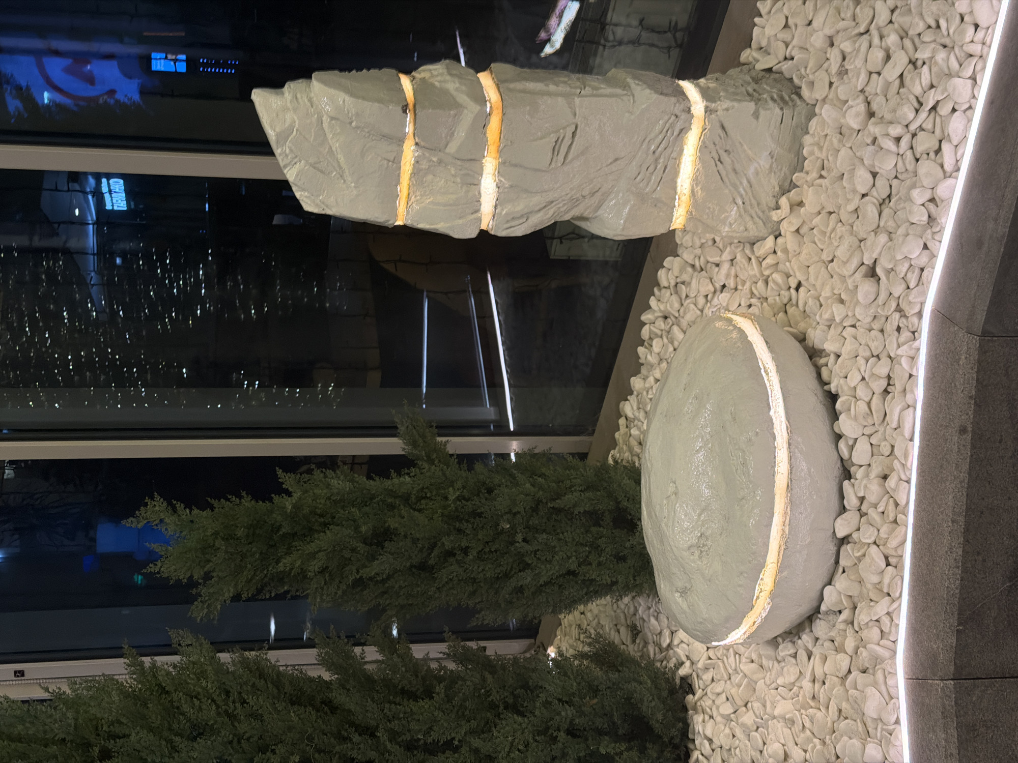 The image shows an indoor decorative arrangement in what appears to be a public space or building interior, potentially in Moscow, Russia, based on the reflections of signage visible in the window. The arrangement consists of:
1. **Decorative Rocks:** Two artificial rocks, one long and segmented and another roughly oval, painted in a light gray color. Both have internal lighting visible through breaks in their structure.
2. **White Pebbles:** The rocks are surrounded by a bed of small, white pebbles.
3. **Greenery:** Part of a coniferous shrub or small tree is visible at the bottom of the image.
4. **Window:** Behind the arrangement is a large window reflecting the cityscape at night.
5. **Edge:** Part of a wall edge is on the right side of the frame.