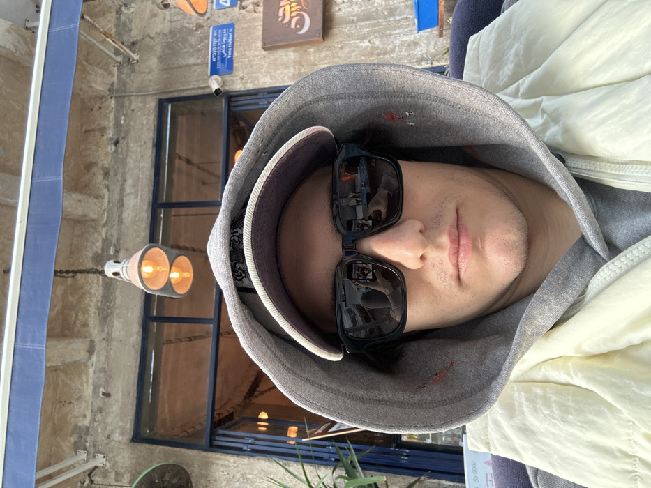 A male individual is depicted in a close-up selfie, seated outdoors on Yona HaNavi Street in Tel-Aviv, Israel. He wears a grey hooded sweatshirt with the hood up, a dark baseball cap, and dark-framed sunglasses that reflect a smartphone held by a hand, indicating he is taking the photo. A light-colored puffer vest is worn over the sweatshirt, featuring a small red embroidered detail on the hood's right side. The background consists of a weathered building facade with a blue awning and a glass-paned entrance. Two industrial-style pendant lights hang from above. A street sign visible on the wall identifies the location as "Yona HaNavi St." in Hebrew and English, positioned below a security camera. To the right, a sign with Arabic script is present on the wall and a blue container is on a table. A green plant is partially visible on the left. The reflections in the sunglasses show a street scene with a parked vehicle.