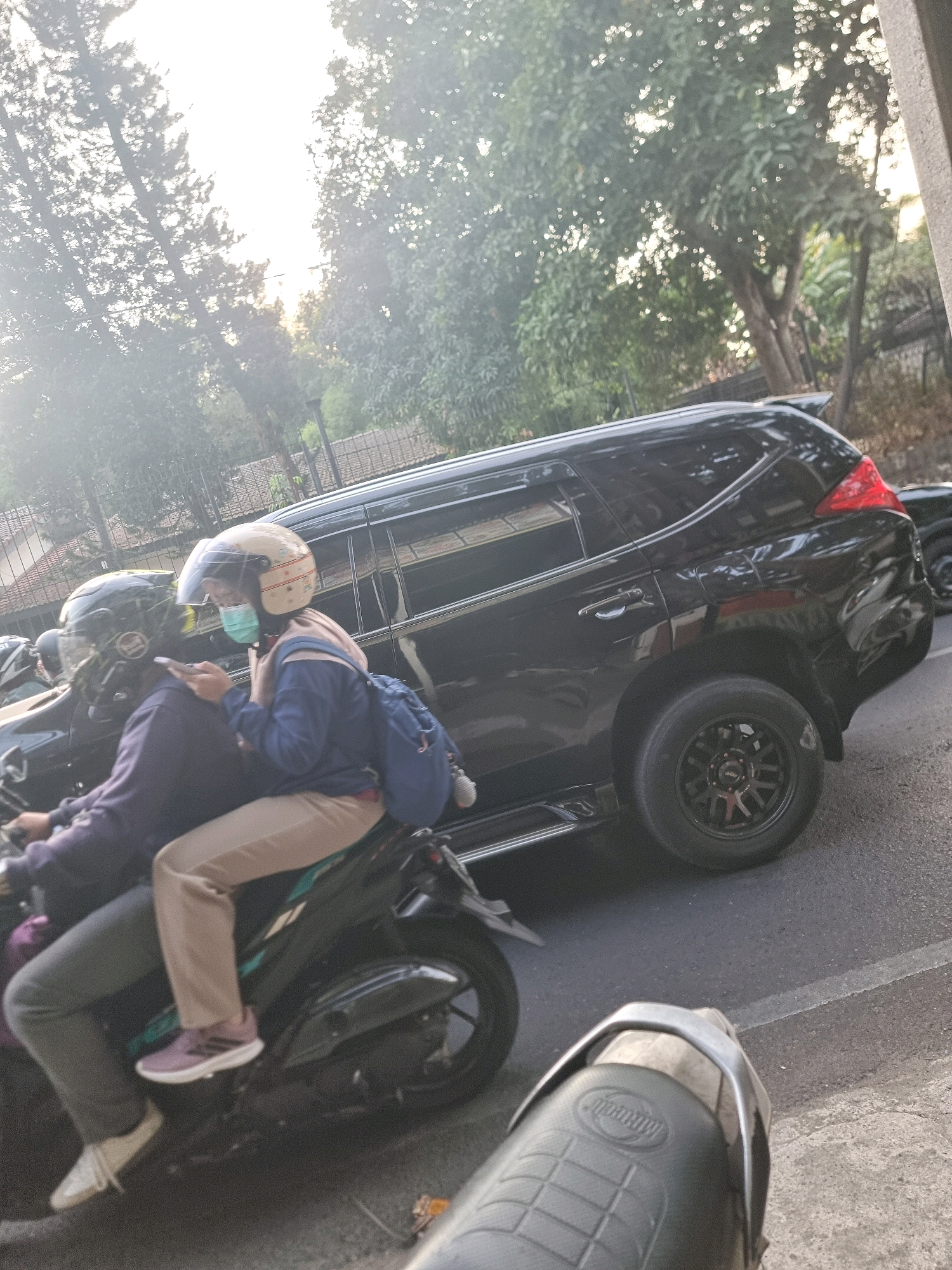In South Jakarta, Indonesia, two individuals are riding a motorcycle. The person in the back is wearing a light-colored helmet with a face shield and a light blue jacket, tan pants, and pink sneakers. They are holding a smartphone and looking at it. The person in the front is wearing a dark helmet with a face shield and a dark purple jacket, and grey pants. They are driving the motorcycle. Behind them, a black SUV is parked on the side of the road, partially obstructing the view of the background which includes trees and buildings. The lighting suggests it is late afternoon.