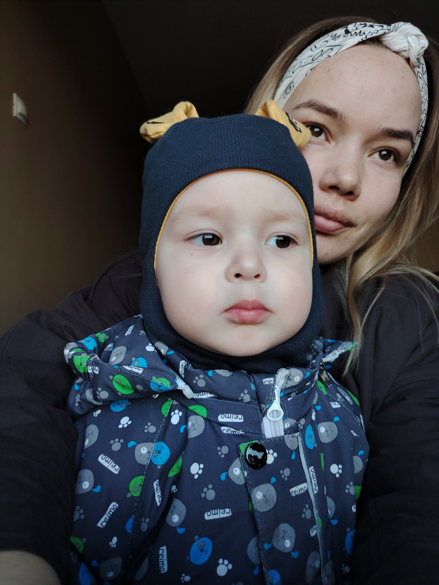 A woman with blonde hair and a white patterned headband holds a young child dressed in a navy blue hooded jacket with a playful dog pattern. The jacket has a white zipper and a black button. The child wears a navy blue knit hat with small yellow appendages resembling ears. The backdrop appears to be a simple interior with a dark wall. The jacket has the brand "reima" printed on it. Location is Марковский, Russia.