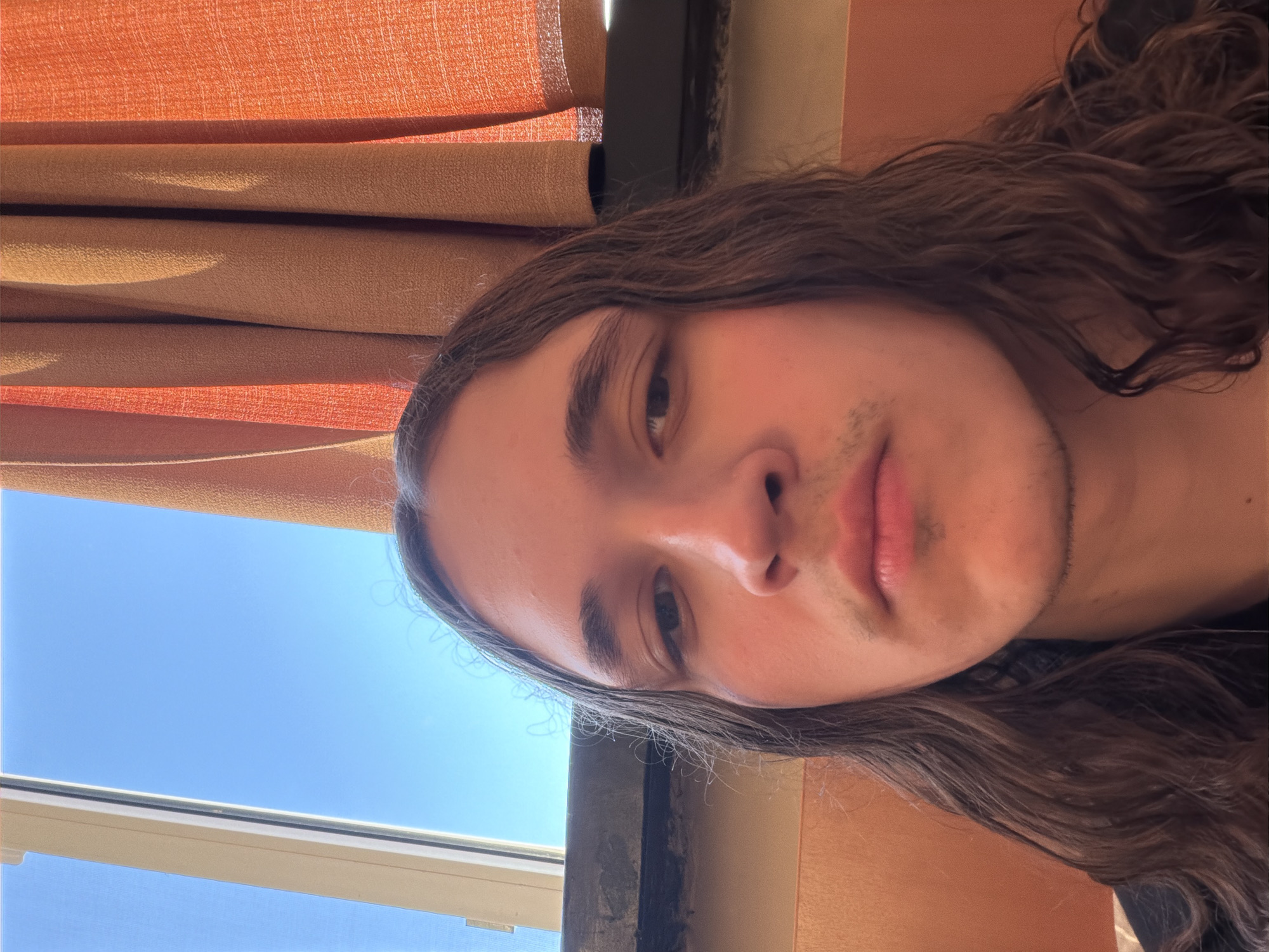 The media file is a portrait-oriented photograph taken indoors near a window in Plovdiv, Bulgaria. The primary subject is a young individual, likely male, with long, dark, wavy hair that frames his face and extends down his shoulders. He has fair skin and displays light stubble around his mouth and chin. His expression is neutral and calm, as he looks directly towards the camera.
The background reveals a combination of indoor and outdoor elements. To the upper left, layered curtains in shades of orange and tan/brown are visible, suggesting an interior setting. A bright, clear blue sky is prominently displayed through a window in the lower left portion of the image, indicating sunny weather during the daytime. A portion of a white window frame is also visible at the bottom edge. The lighting suggests strong natural light coming from outside, illuminating the subject's face. There is no visible text in the image. The overall impression is a casual, sunlit selfie or portrait.