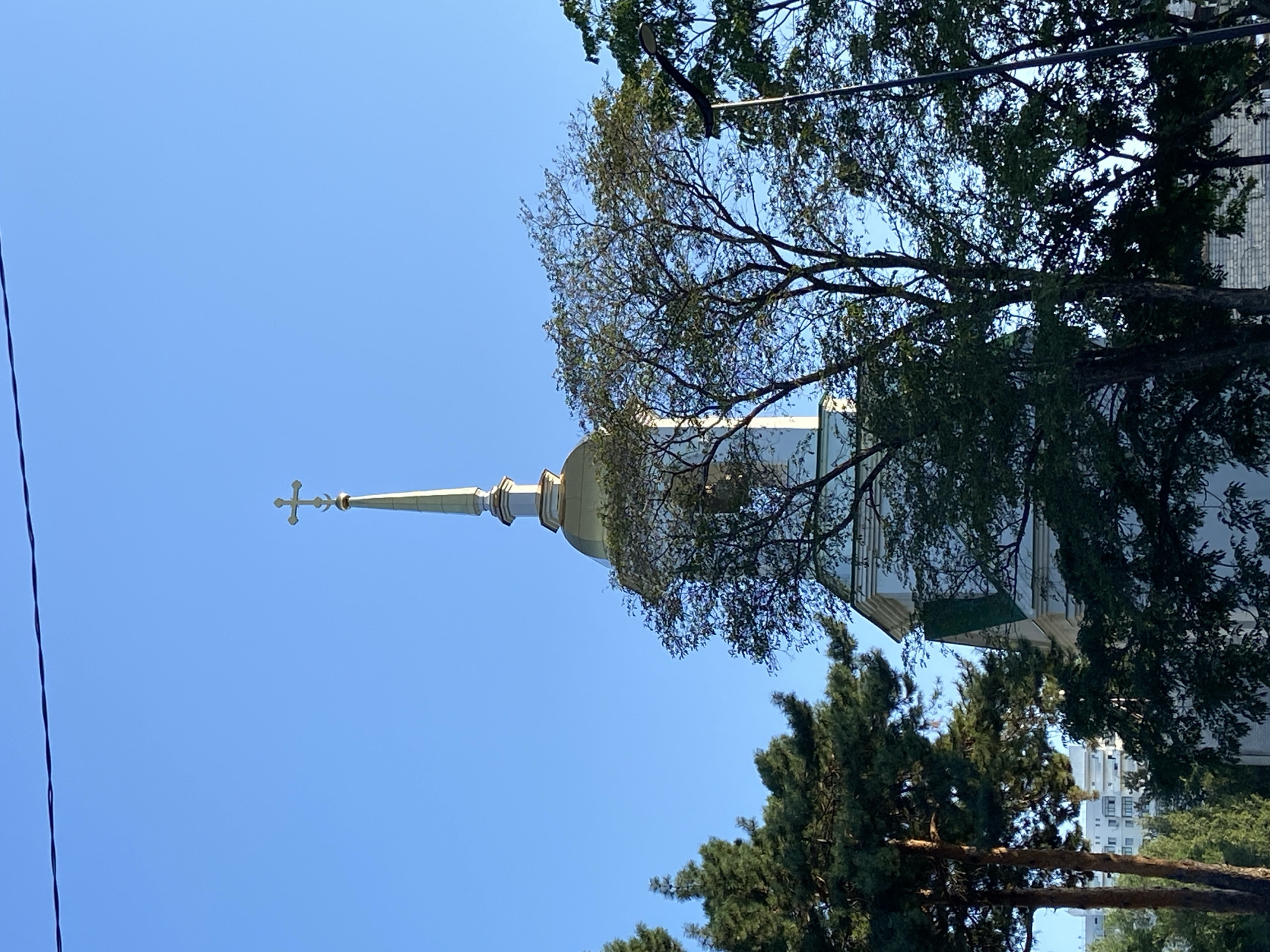 A church steeple topped with a gold cross is visible against a clear blue sky, partially obscured by trees. The church itself has a gold dome and white walls with green trim. In the background, a modern apartment building can be seen. A power line runs horizontally across the top of the frame. The lighting suggests it is daytime, likely morning or late afternoon due to the warm tones. The image appears to be taken from ground level, looking upwards at the structure. This scene is consistent with the architecture found in Blagoveshchensk, Russia, where many Orthodox churches feature similar design elements.
