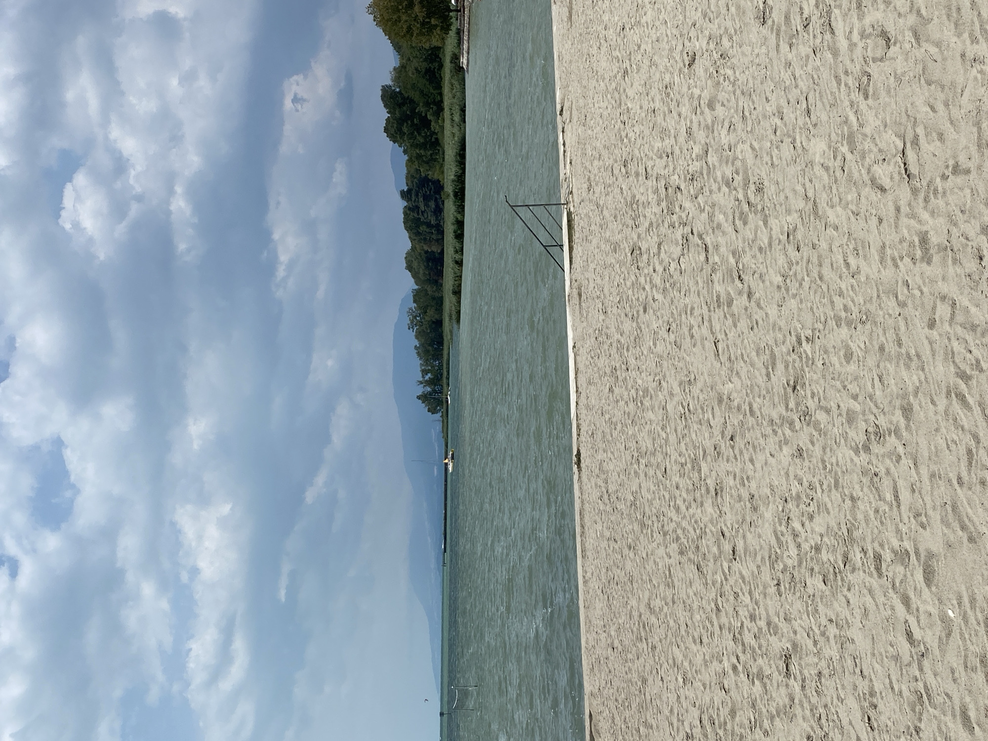 The image depicts a sandy beach leading to a body of water under a cloudy sky. In the foreground, the sand is textured with visible footprints. A short, white concrete barrier separates the beach from the water. A metal railing with several steps is partially submerged in the water. The water is choppy with visible waves. In the middle ground, reeds and trees line the shore. A small orange boat is visible on the water, along with a distant sailboat and a kiteboarder. Mountains form the backdrop. The scene is outdoors and appears to be a public beach.