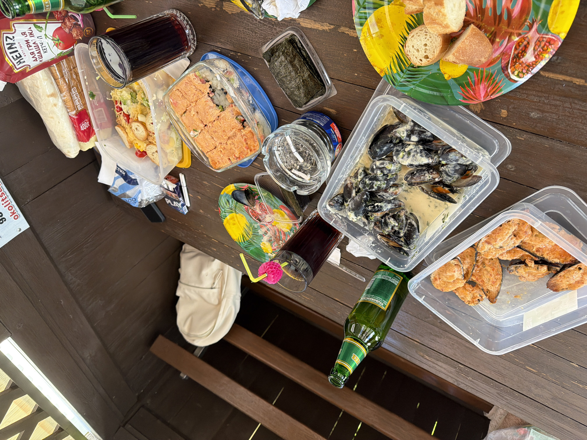 The image depicts an outdoor picnic table laden with food and drinks. In the foreground, a clear plastic container holds a portion of cooked mussels in a creamy sauce. To the right of this, a colorful plate displays slices of bread and what appear to be lemon wedges. Behind the mussels, another clear container holds fried fish or chicken pieces. Further back, a rectangular container holds what seems to be sushi or a similar dish with a reddish topping. A glass of dark liquid with a straw and a pink pom-pom garnish is visible. Adjacent to this is a bottle of green glass beverage, possibly beer. A ketchup bottle with Russian labeling is also present. Several plastic containers and wrappers are scattered across the wooden table, which appears to be located outdoors, likely in a rustic setting given the wooden planks and overhead covering. The overall scene suggests a casual meal being enjoyed outdoors. The location is identified as Staroderevyankovskaya, Russia, suggesting a rural or semi-rural setting.