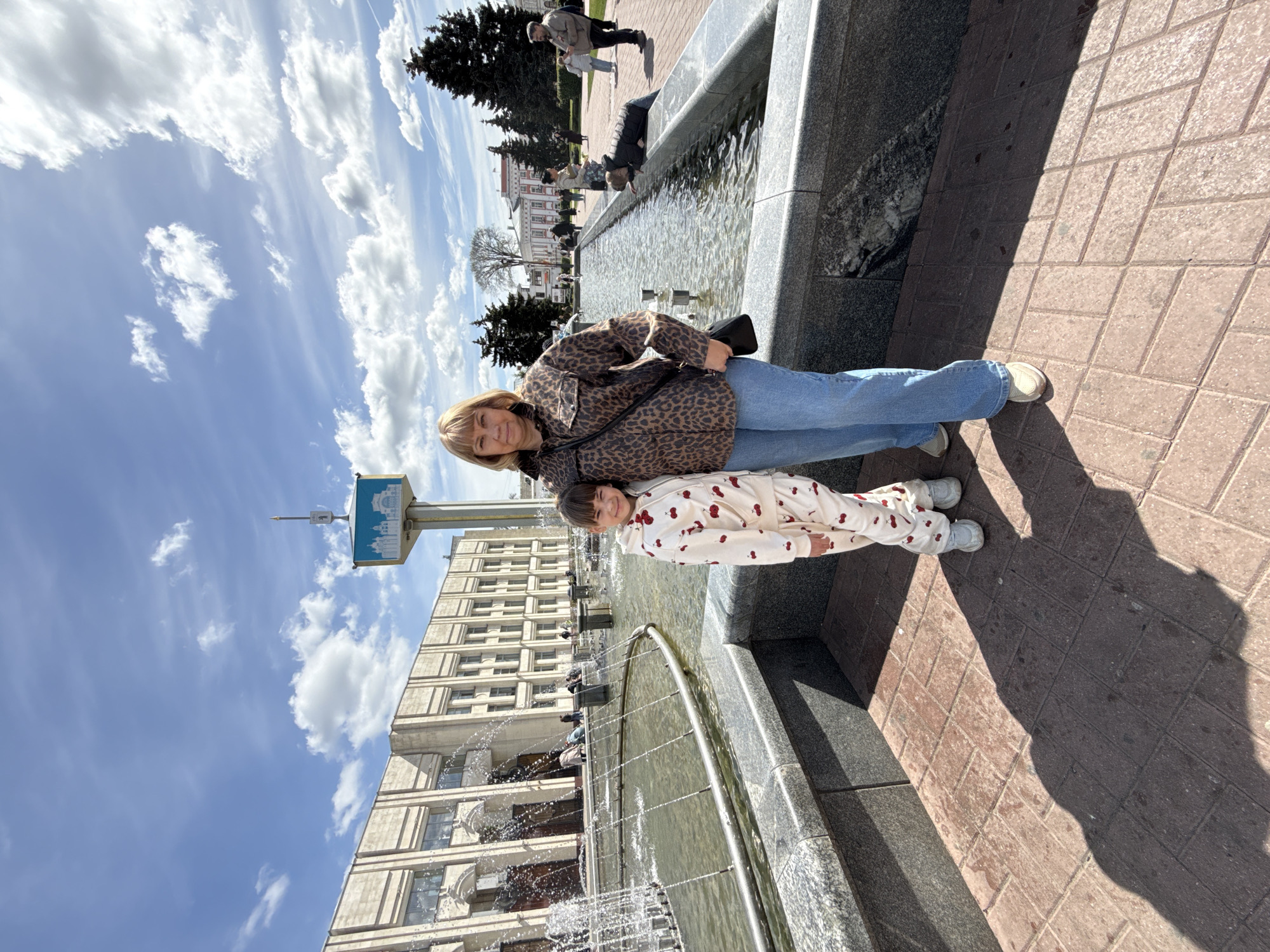 A medium shot of a woman and a child posing in front of a fountain in Unknown, Unknown. The woman is standing and wearing a leopard-print jacket and jeans. The child is wearing a white pajama set with red cherries printed on it. They are standing next to the fountain which is made of gray stone and has a metal railing around it. There is a building in the background with a tall tower with a sign on top. The sky is blue with white clouds. The ground is paved with red bricks.