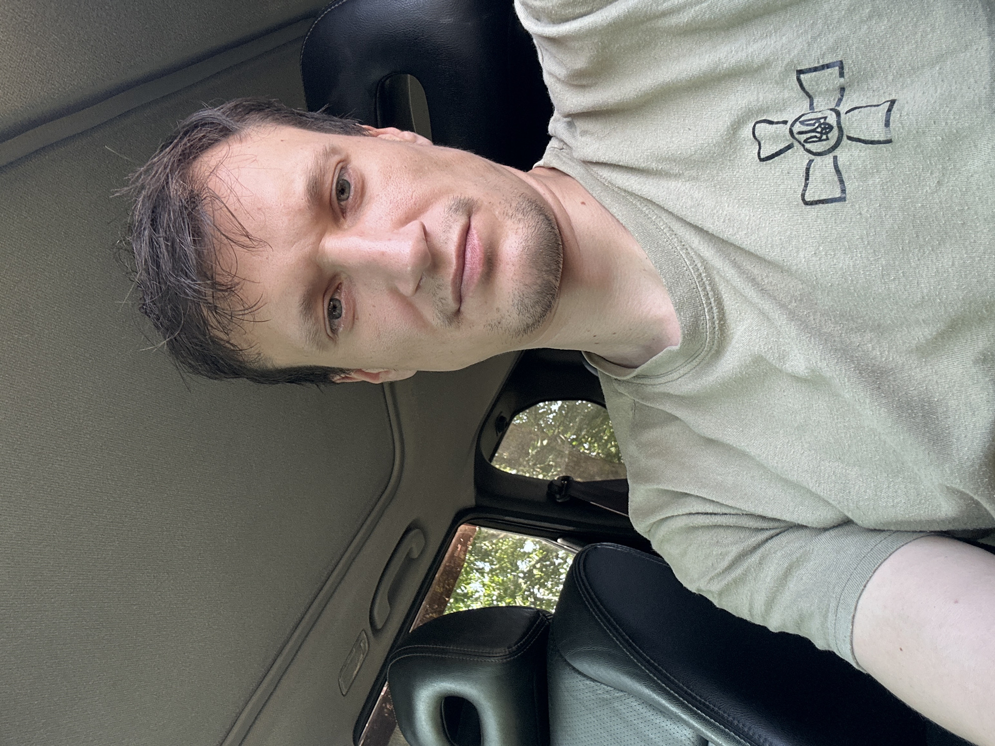 A man is sitting in the driver's seat of a car. He has short, dark hair and is wearing a light-colored t-shirt with a black symbol on the left chest. The symbol appears to be a cross with a coat of arms in the center. The interior of the car is visible, including the dashboard, headrests, and a sunroof. Sunlight streams in through the windows, illuminating the interior and highlighting the textures of the seats and the headliner. Outside the car, trees and foliage are visible, suggesting a natural or suburban setting. The image was taken from a slightly elevated angle, looking down at the man.