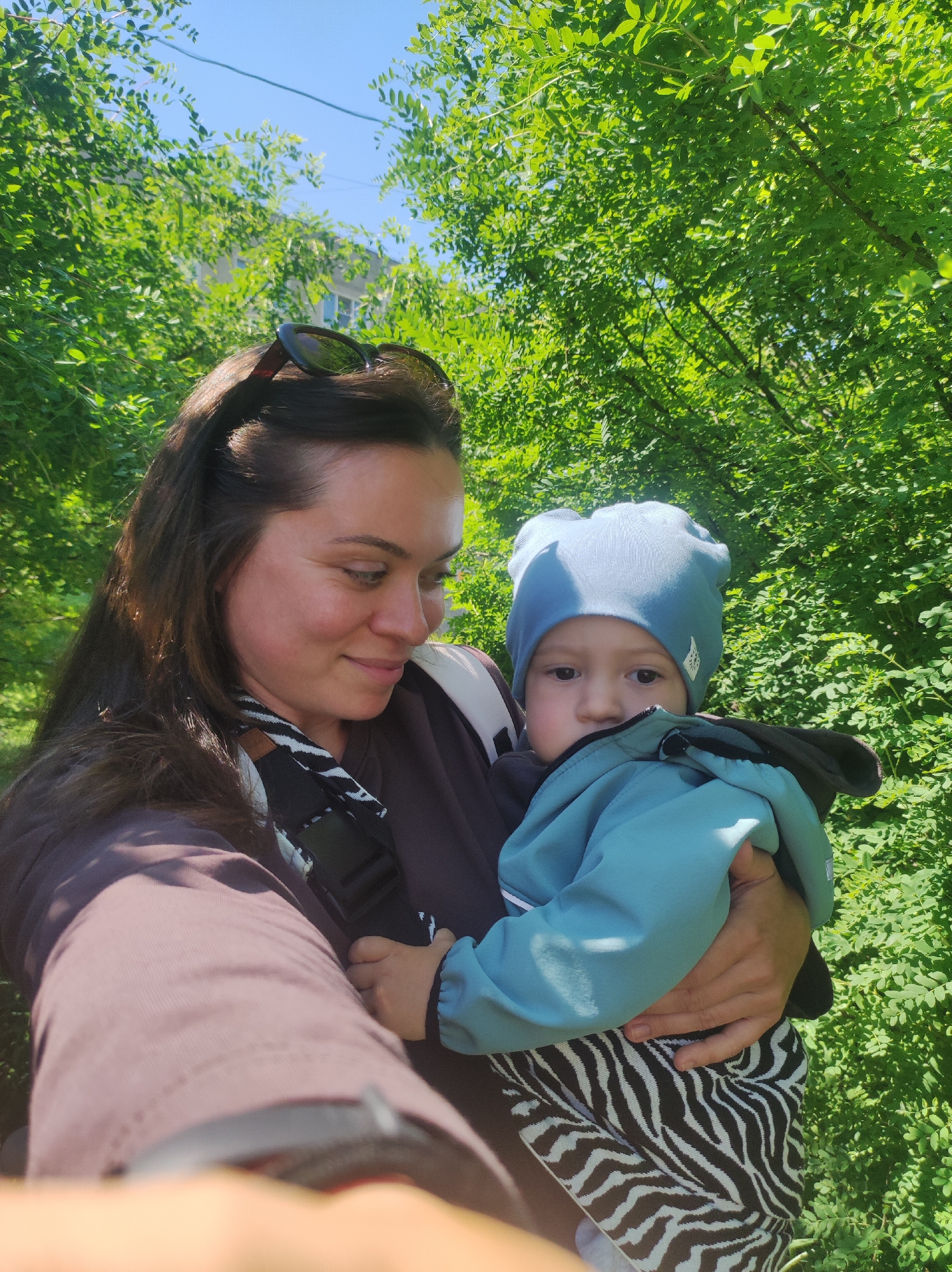 This outdoor portrait features a woman holding a young child against a backdrop of lush green foliage and a clear blue sky, in Chaikovsky, Russia. The bright sunlight suggests it is daytime.
The woman, with long, dark brown hair and sunglasses perched on her head, occupies the left side of the frame. She wears a dark brown long-sleeved top and a black and white patterned strap, possibly from a carrier, across her shoulder. Her expression is a soft, affectionate smile as she looks down at the child in her arms.
The child, held securely by the woman, is positioned on the right. They are dressed in a light blue beanie with subtle ear-like protrusions and a small, white, illegible emblem. A blue-teal jacket covers their upper body, with the collar slightly obscuring their chin. The child's dark, observant eyes are visible, looking slightly off-camera. Below the jacket, the child's attire features a distinct black and white zebra print.
The background is dominated by dense, vibrant green leaves from trees and bushes, indicating a natural setting like a park or garden. A small portion of a light-colored building can be seen through the foliage in the upper left, beneath a bright, cloudless blue sky. The overall impression is one of warmth and maternal affection.