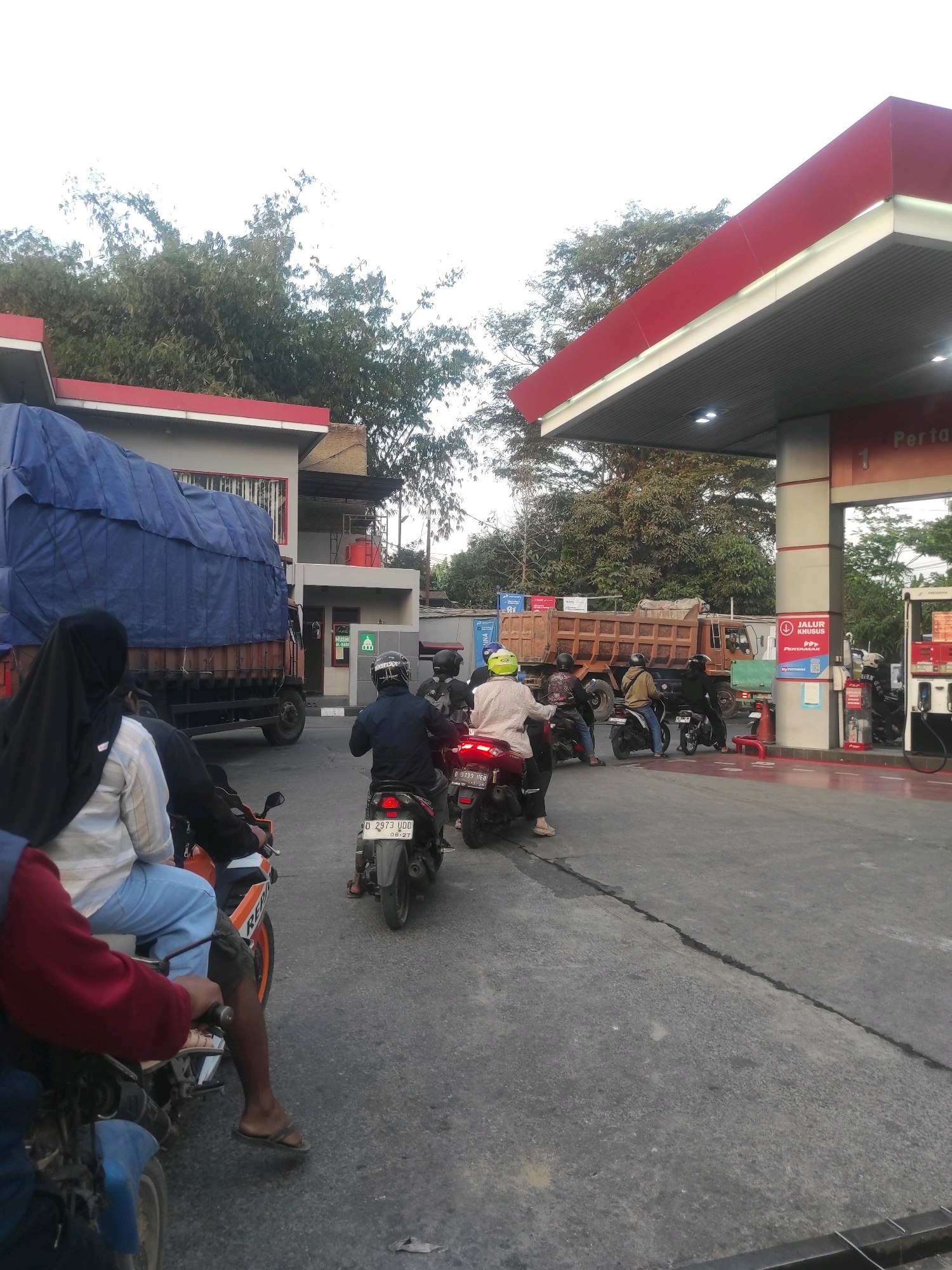 At a gas station in Padalarang, Indonesia, several people on motorcycles are stopped. The gas station has a red and white awning supported by columns, and a Pertamina logo is visible. A large truck covered with a blue tarp is parked on the left side of the image, next to a building. In the foreground, a person wearing a maroon jacket and blue pants is seated on a motorcycle. Behind them, other motorcyclists are also present. Trees and vegetation surround the gas station. The sky is overcast.