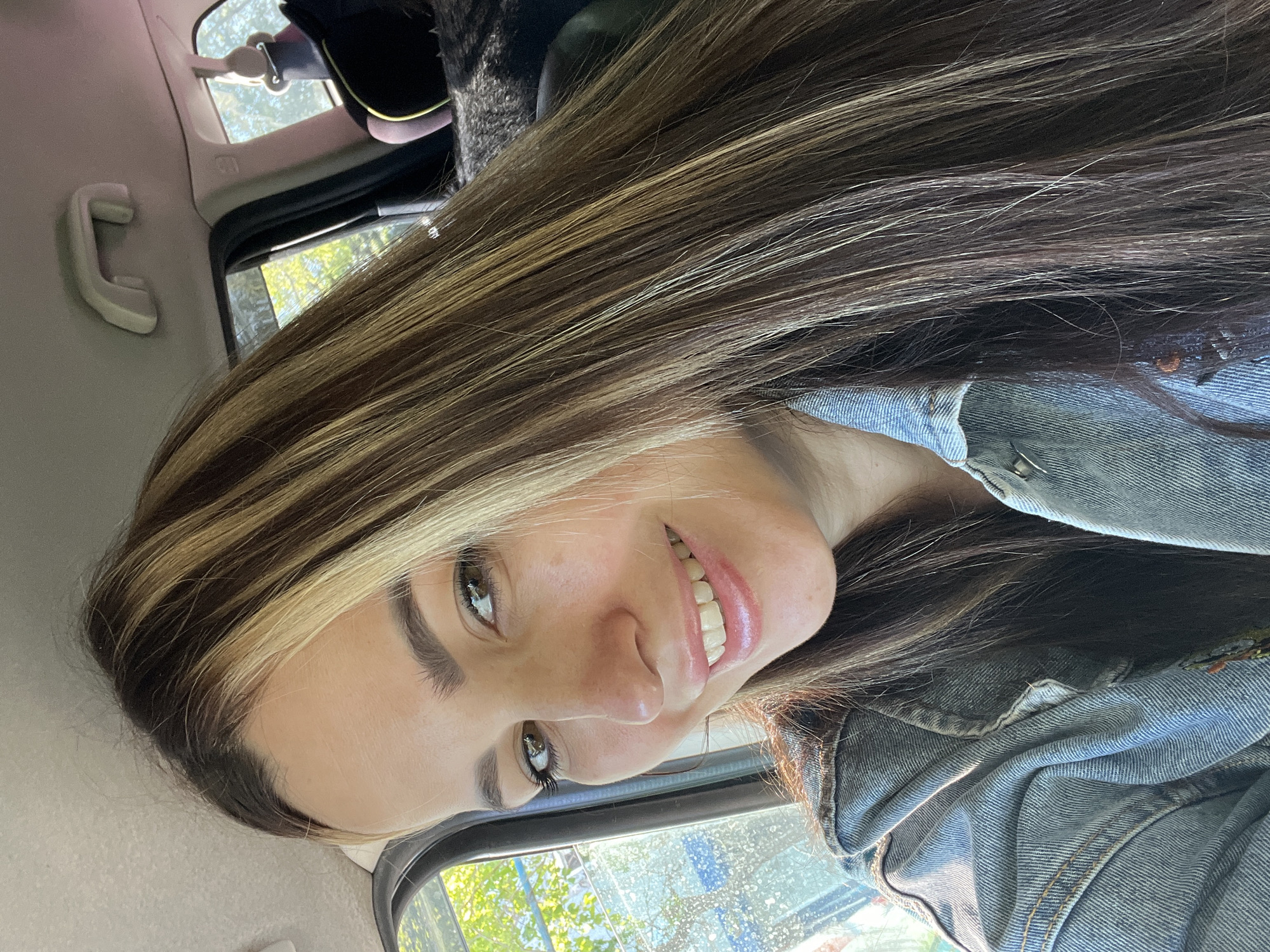 A woman with brown and blonde hair, wearing a denim jacket, is smiling and looking at the camera from the inside of a car. The car interior is visible, including the window, handle, and ceiling. The location is unknown.