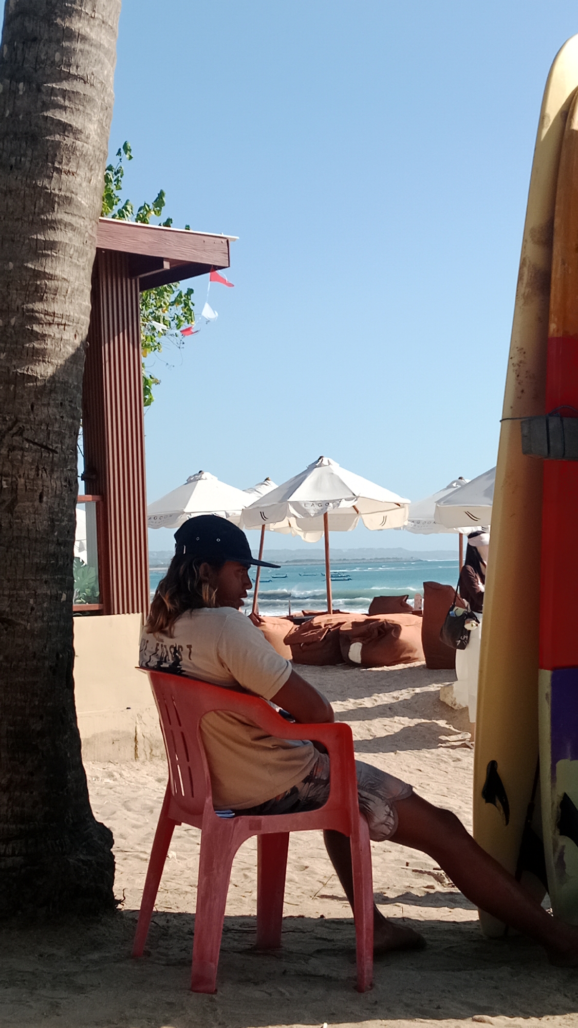 A man wearing a black baseball cap and a tan t-shirt sits on a red plastic chair facing right. He has long, light brown hair and is wearing patterned shorts. He is sitting on a sandy beach, with a palm tree to his left and a surfboard to his right. In the background, there are white umbrellas, beanbag chairs, and the ocean with several boats visible. The scene appears to be in Kuta, Indonesia, based on the context provided. The sky is clear and blue.